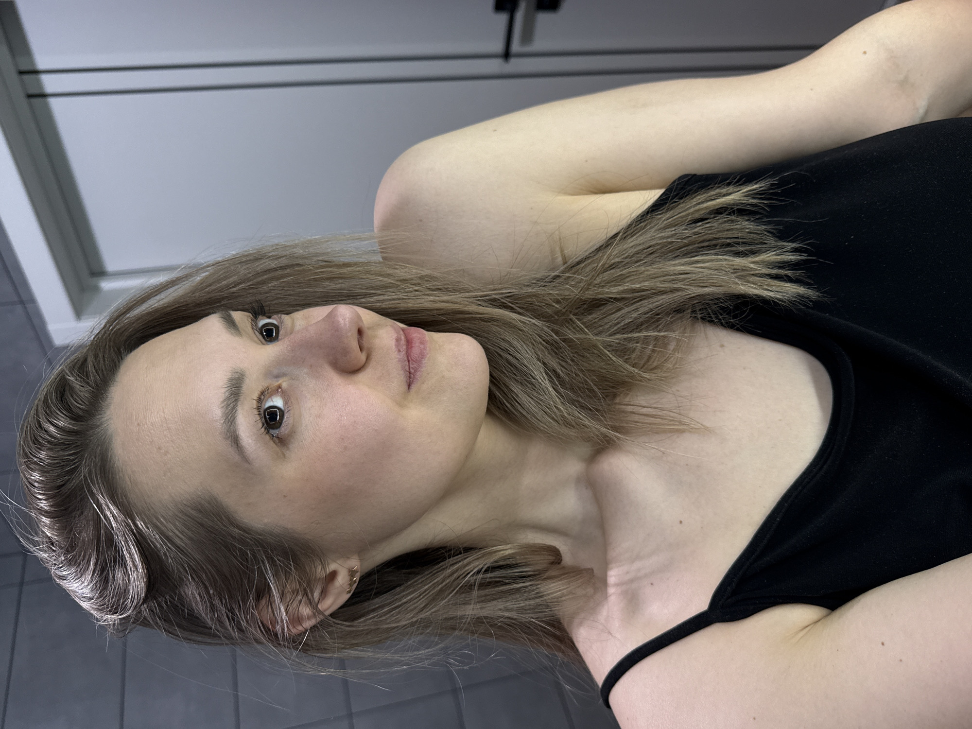 A woman with fair skin and light brown hair is posing for a close-up shot. She's wearing a black tank top. The background consists of a white cabinet or wall with dark handles and a gray tiled floor. The location in the city of Chaikovsky, Russia, is not discernible from the image.