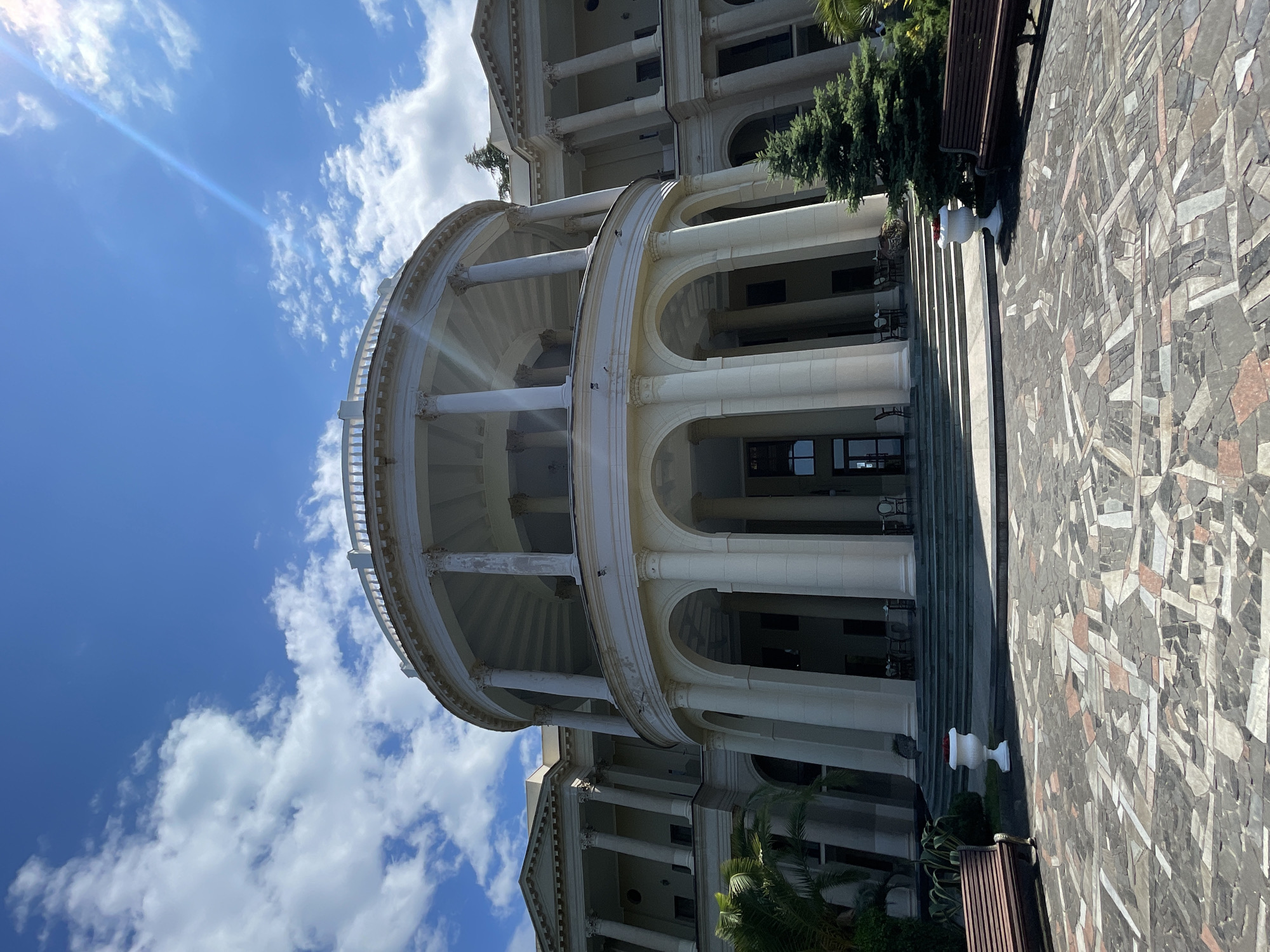 The image shows the grand, white, neoclassical facade of the Sanatorium Primorye in Gagra, Abkhazia. The building features a prominent semi-circular colonnade on the ground floor, with large arches and imposing columns leading up to a second level. Above this, a curved balcony or walkway is supported by a ring of slimmer columns. The facade is adorned with more classical architectural elements, including pediments and pilasters.
In the foreground, a cobblestone courtyard with a mosaic pattern of various colored stones stretches out. Two dark wooden benches are placed on either side of the courtyard. A large, white decorative urn sits near the steps leading up to the building on the left. A small, well-maintained evergreen tree stands to the right of the main entrance. The sky above is a bright blue, scattered with fluffy white clouds, and a bright sunbeam creates a lens flare effect. The overall impression is one of grandeur and historical significance, set against a clear, sunny day.