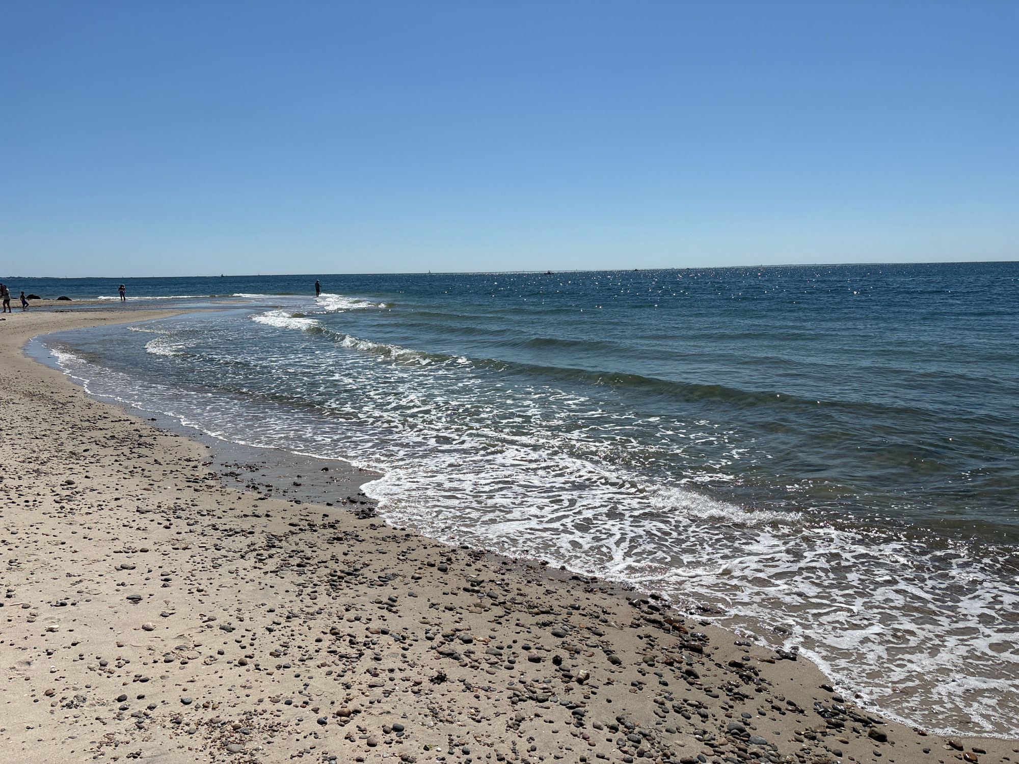The image depicts a serene beach scene on a clear, sunny day. The foreground is dominated by a sandy beach covered with small pebbles and shells. Gentle waves with white foam are lapping at the shore. In the mid-ground, several individuals are visible along the water's edge and in the shallow water, appearing to be enjoying a leisurely day. A few sailboats and other vessels are spotted on the horizon, dotting the vast expanse of the deep blue ocean. The sky is a clear, brilliant blue with no clouds. This scene is likely located at a beach in Waterford, United States of America.
