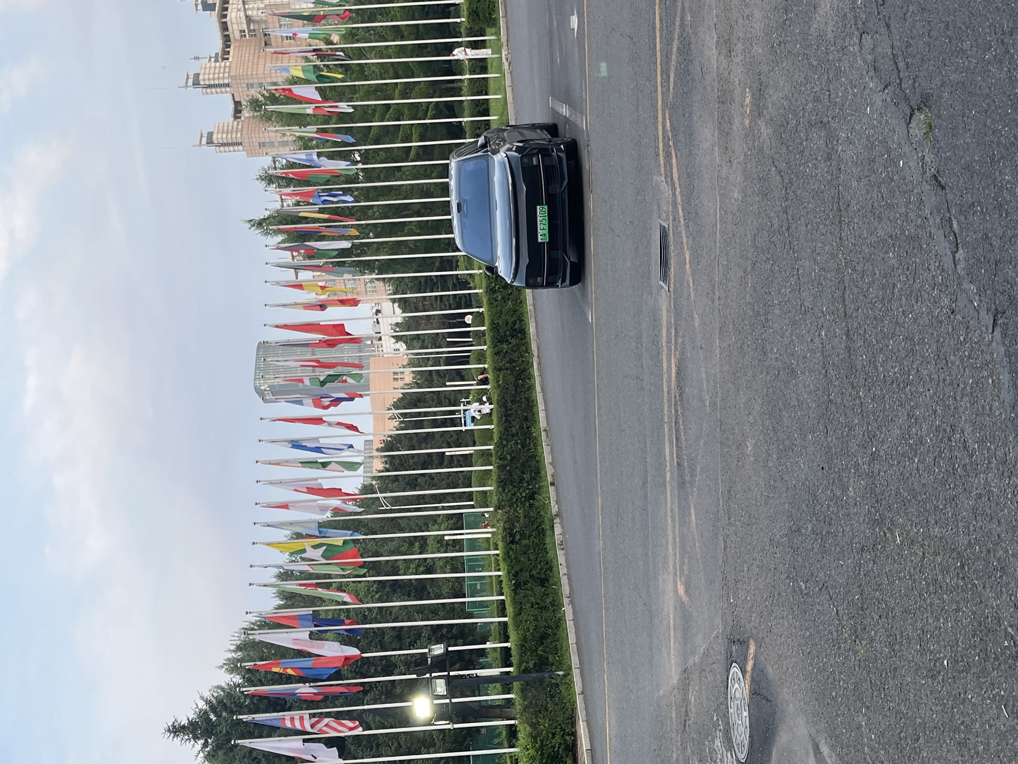 A black sedan with a green license plate is parked on an asphalt surface in front of a row of international flags. The flags are on white poles and are displayed against a backdrop of lush green trees and modern buildings. The sky is overcast. The license plate has the characters "A F75109". The scene appears to be in a city, possibly a diplomatic or international affairs related location. Based on the license plate format and design, it is likely in Changchun City, China. There are a few people in the background, including one person in a white shirt standing near the flags. The asphalt surface shows some wear and tear, with cracks visible. A manhole cover is also present on the asphalt.