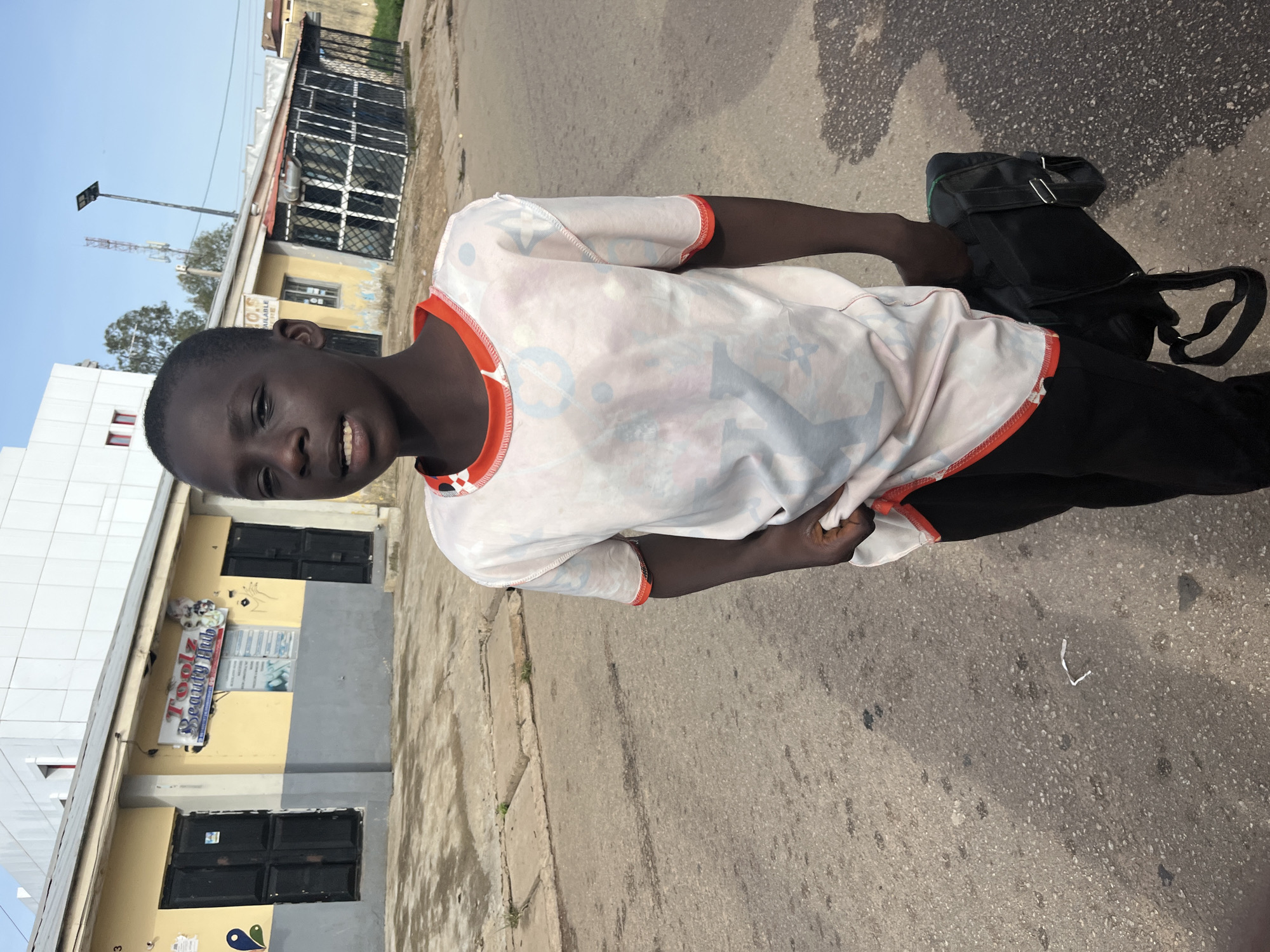 A young boy stands on a street in Jos, Nigeria, wearing a light-colored t-shirt with a repeating pattern and black pants. He is holding a black bag in his right hand. In the background, a yellow and gray building with dark doors and signage is visible. The street is paved with asphalt, and there are some concrete slabs to the left. The sky is clear and blue.