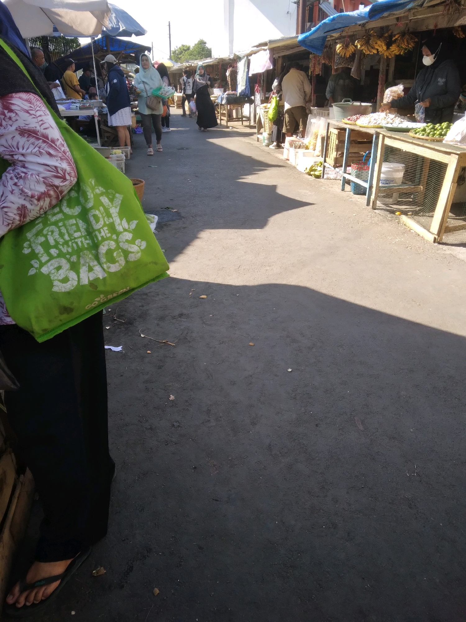 The image depicts a bustling outdoor market scene in Sokaraja, Indonesia. A person in the foreground, wearing black pants and flip-flops, is holding a large, bright green reusable shopping bag with white text that reads "ECO FRIENDLY WITH THE BAGS" and "GO GREEN". Behind them, other market-goers are visible browsing various stalls. One stall displays an abundance of bananas hanging overhead, and another features piles of produce, including onions and limes, on wooden tables. A vendor, wearing a face mask, stands behind one of the stalls. The ground is paved with asphalt, and sunlight casts strong shadows across the scene, indicating it is daytime. The overall impression is one of a lively local market.