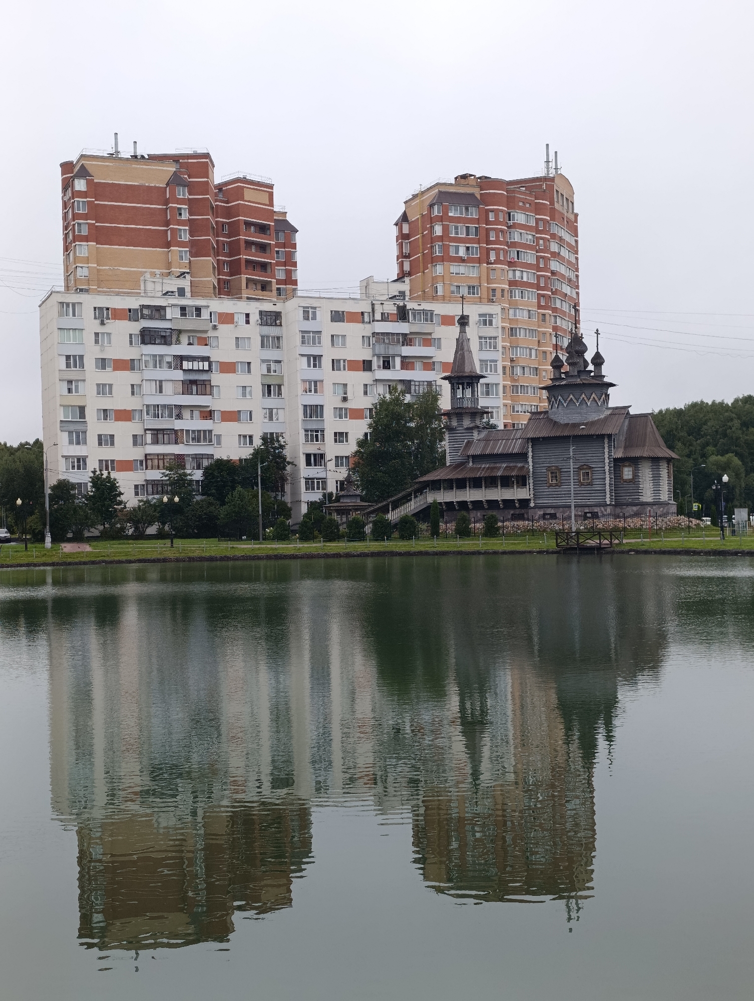 This is a wide, overcast, eye-level outdoor shot of a tranquil pond and its reflection, with a large residential building complex and a wooden church visible in the background. The pond is in the foreground, taking up roughly the bottom half of the frame, and its surface is calm, creating a clear reflection of the scene above. The reflection shows the multi-story residential buildings, the church, trees, and the cloudy sky.
In the middle ground, on the far side of the pond, stands a large, modern residential building complex. It appears to be composed of at least two connected towers. The tower on the left is shorter and has a facade primarily of light beige brick with darker red brick accents on its upper levels. The tower on the right is taller and more curved, with a similar brick pattern of alternating beige and red sections, interspersed with numerous windows. In front of these towers, and slightly to the right, is a traditional-looking wooden church. The church is grey, made of logs, and features multiple onion domes, a bell tower, and a covered walkway. A staircase leads up to the church's entrance.
Behind the church and extending to the right are dense green trees, forming a natural backdrop. To the left of the residential complex, there are also trees and some signs of a street or pathway with lampposts. The sky is uniformly grey, indicating an overcast day. The overall impression is a juxtaposition of modern urban architecture with historical religious structures, set against a natural element of water. The location is identified as Kiyevsky, Russia.
