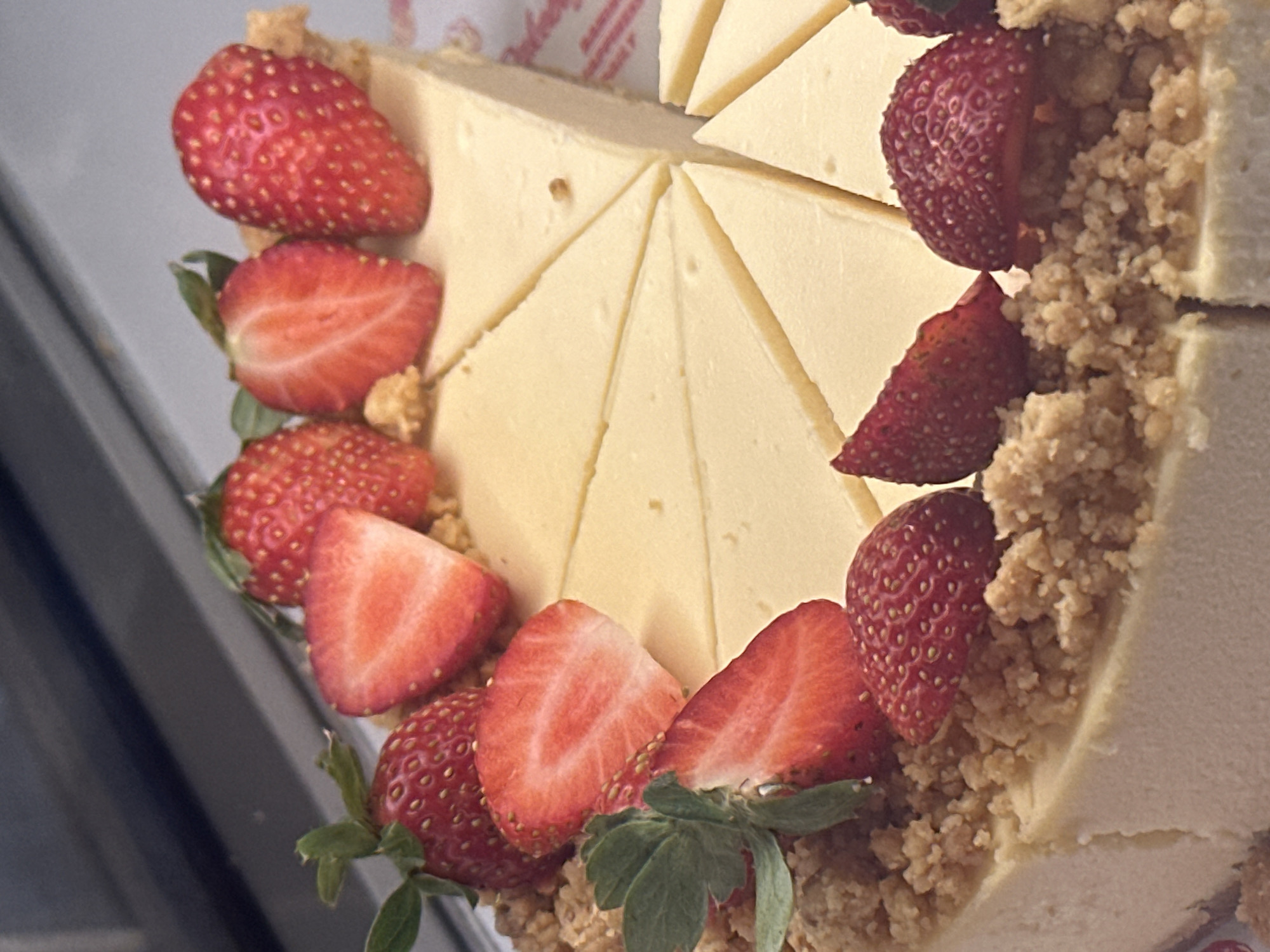 A whole cheesecake is presented in a close-up shot, showcasing its creamy texture and tart topping. The cheesecake has been sliced into several triangular portions, revealing the smooth, pale yellow interior. It is adorned with fresh strawberries, some whole and some halved, scattered across the top. Crumbled pieces of what appears to be cookie or pastry are sprinkled around the strawberries, adding a rustic and appealing finish. The cheesecake rests on a white plate with a red patterned border visible on the right side. The background is blurred, suggesting an indoor setting, possibly a bakery or a restaurant display, with hints of a grey surface and a metallic edge in the upper portion of the image. There is no indication of specific locations within Bac Nha Trang Ward, Vietnam.