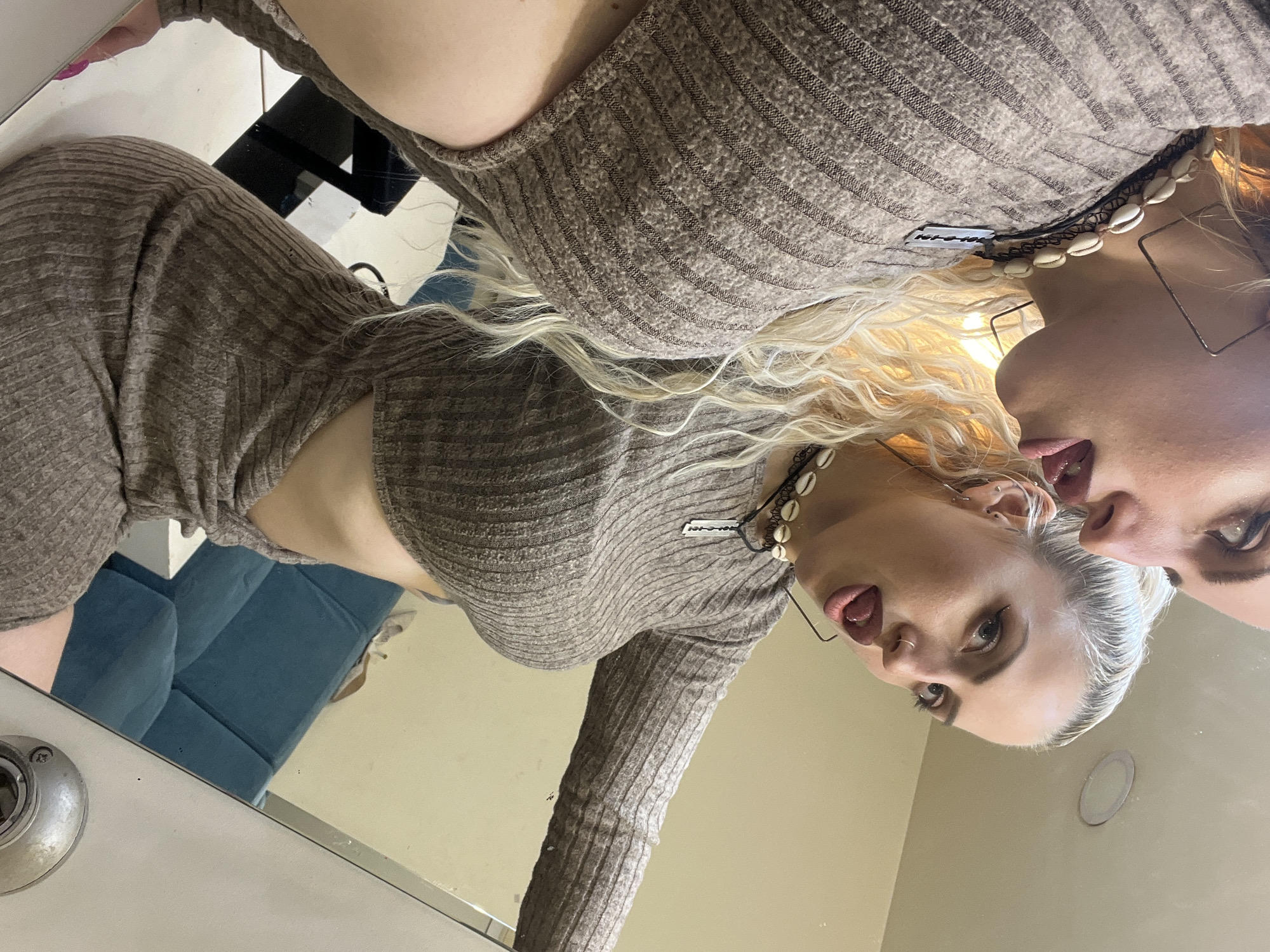 A selfie of a woman reflected in a mirror. She is wearing a ribbed brown outfit and a shell necklace. Her hair is blond and white. A blue sofa is visible in the mirror. The location is not discernible as Plovdiv, Bulgaria.