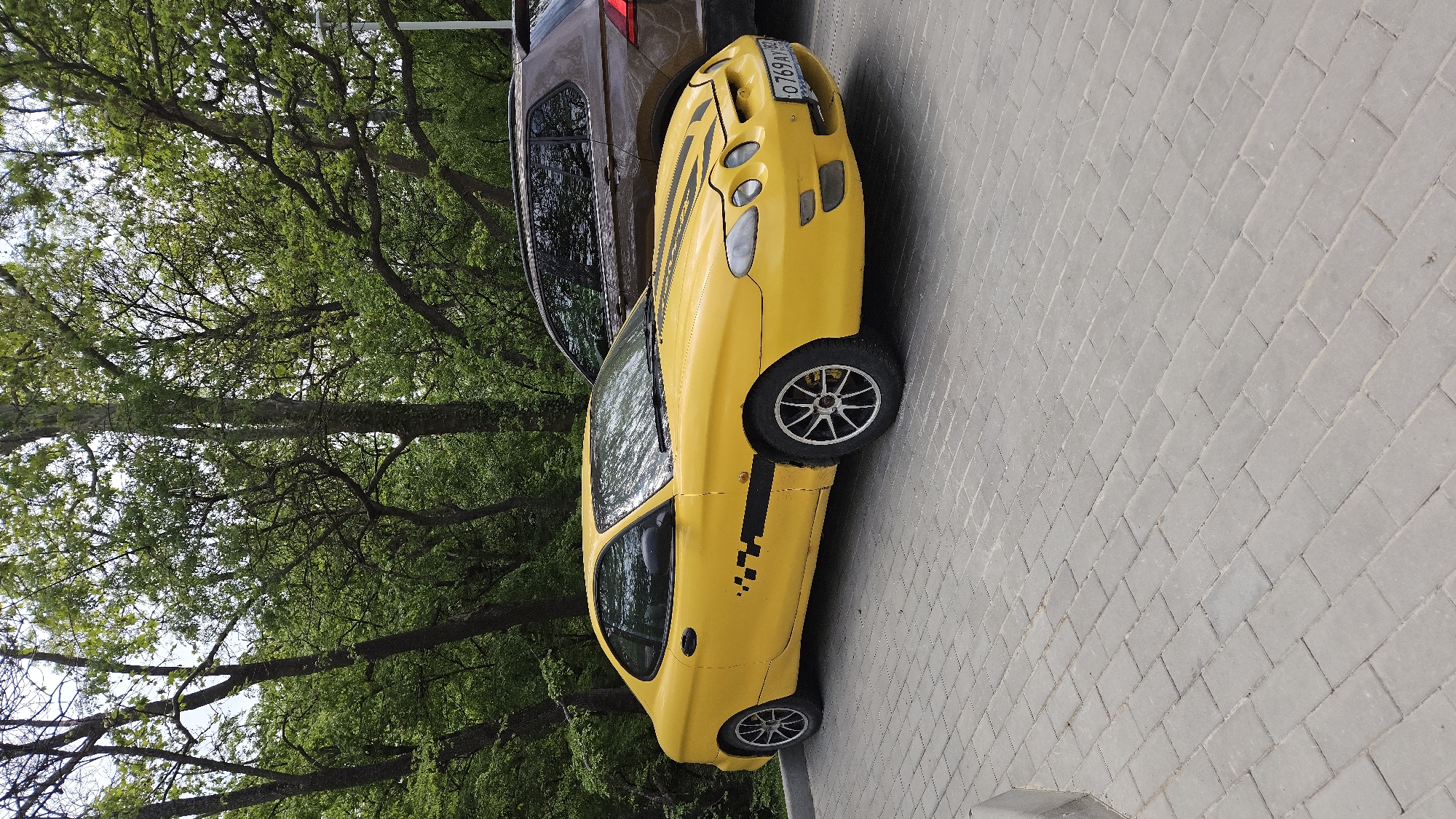 The image shows a yellow car with black stripes and a taxi logo parked on a paved surface. A brown SUV is parked next to it. Trees with green leaves are visible in the background. The location is in an unknown city.