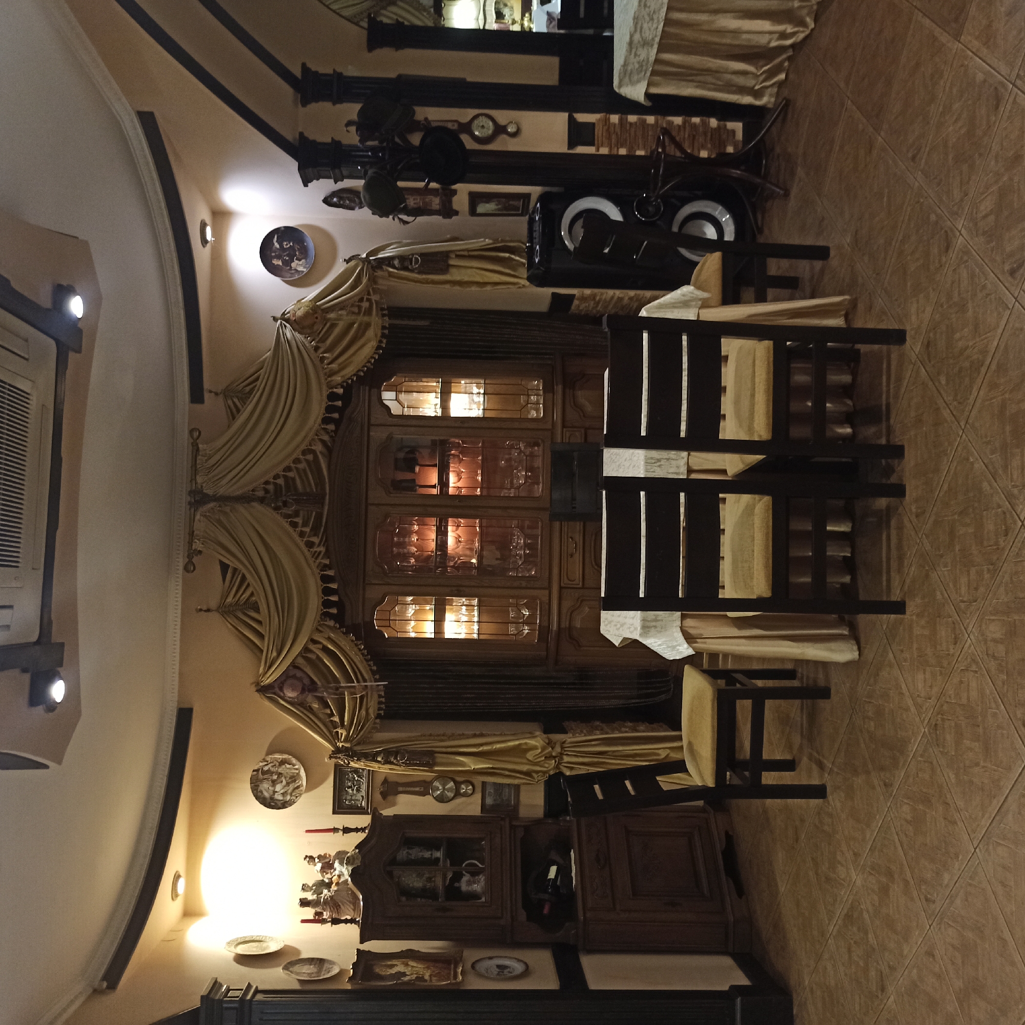 The image depicts the interior of a restaurant or dining hall, possibly located in Chernivtsi, Ukraine, judging by the style and décor often found in establishments in the region. The scene is dominated by a large, ornate wooden hutch filled with glassware and illuminated from within. In front of the hutch, a dining table is set with a white tablecloth and is surrounded by dark wooden chairs with yellow upholstered seats. The room features warm, ambient lighting, with recessed spotlights on the ceiling and decorative wall lights. The walls are painted a pale yellow or beige, and the floor is tiled with brown, square tiles.
To the left of the hutch is another wooden cabinet, also displaying decorative items and possibly serving as a display piece. On the wall above this cabinet, several decorative plates are mounted. To the right of the hutch, a large black speaker is visible, along with a decorative coat rack with several hats. The room is draped with golden curtains, adding to the overall opulent and slightly traditional ambiance. The arrangement suggests a formal dining setting, possibly for a restaurant or a private dining room. There are no people visible in the image. The scene is static and appears to be set up for patrons, but is currently empty.