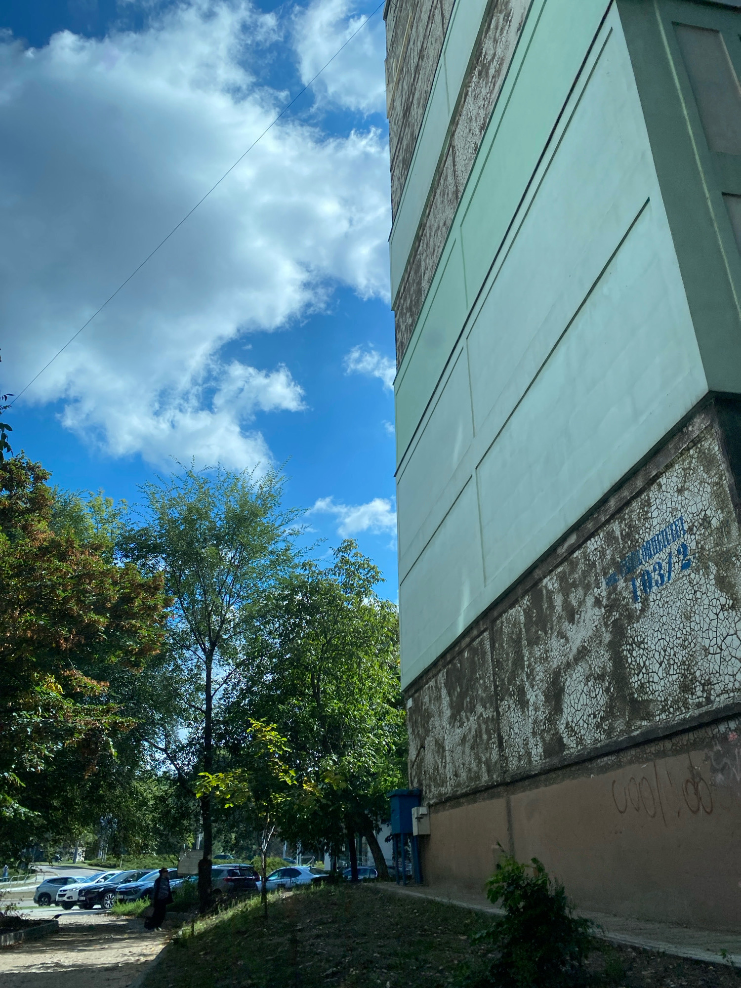 An exterior, eye-level, slightly tilted-up, medium shot captures the side of a building with a bright blue sky and clouds in the background. The building, painted in light green and beige, features several horizontal divisions. The lower portion of the building exhibits signs of wear and graffiti, with a faded blue number "1031/2" visible. Green trees with some autumnal foliage frame the scene on the left, and a paved area with parked cars and a few individuals is visible in the lower left corner. The overall environment suggests an urban setting, possibly in Chișinău, Moldova, characterized by residential buildings and greenery.