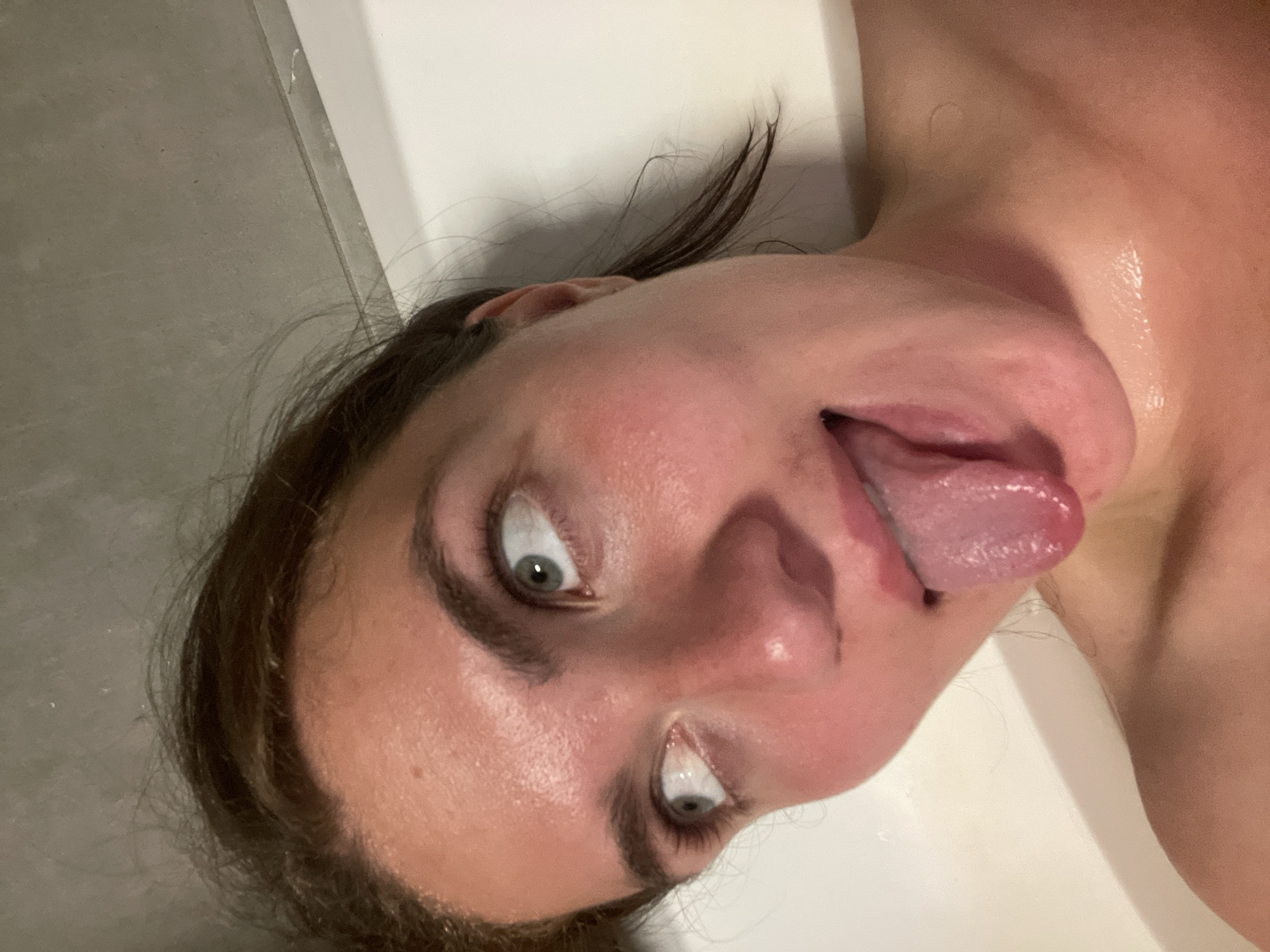A close-up, slightly angled view of a person's face, likely female, with light skin. The individual has dark hair and light-colored eyes, and they are sticking their tongue out. The skin appears to be moist, perhaps from being in a humid environment. In the background, there are elements suggesting a bathroom setting, with gray tiles on the left and a white surface. Мосметаллоконструкция Онт, Russia is not visually identifiable in the image.