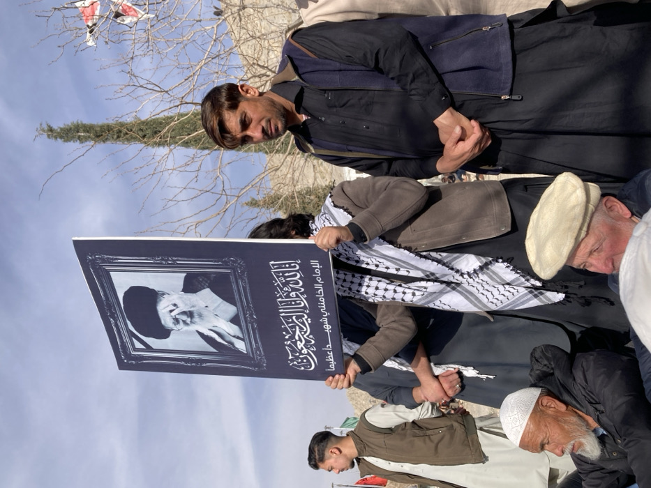 An outdoor scene in Quetta City Tehsil, Pakistan, depicts a gathering of individuals under a clear sky. In the central foreground, a person holds a large black rectangular sign featuring a white-framed black and white portrait of an elderly bearded man in a black turban, identified as Ayatollah Ruhollah Khomeini. Below the portrait, white Arabic script reads, "Indeed, to Allah we belong and to Him we shall return," followed by "Imam Khamenei, a great martyr." The sign-holder is partially obscured, wearing a brown jacket and a black and white checkered keffiyeh.
To the right of the sign, a man with dark hair and a light streak wears a black shirt and a dark blue sleeveless vest, with his hands clasped. To the left, a young man in a light brown vest over light traditional attire looks to his right. In the lower left foreground, an elderly man with a white beard and white prayer cap sits, looking left. Another elderly man with a light Pakol cap is partially visible in the lower right foreground.
The background shows sparse, bare trees and a tall, slender coniferous tree. Distant mountains or hills are visible, and faint flags, including red and green ones, are partially discernible on the left. The ground appears to be unpaved. The individuals are engaged in a collective observation, suggesting a commemoration or public assembly.