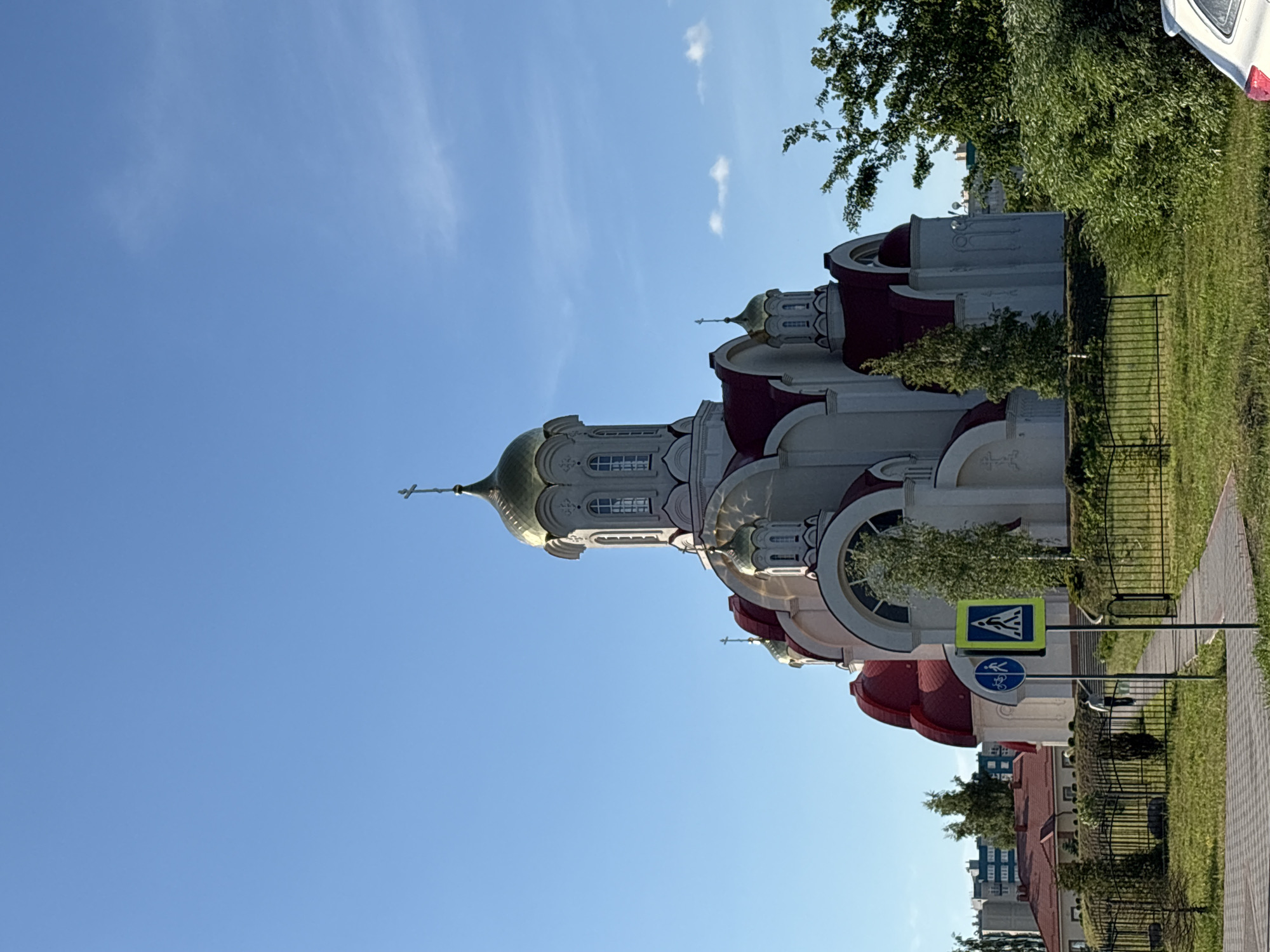 The image captures a prominent Russian Orthodox church under a bright, clear blue sky with a few wispy clouds, indicating a sunny day. The church features pristine white walls, distinctive red roofs, and multiple gilded onion domes, each topped with a cross. Architectural details like arched windows, decorative cornices, and multi-tiered structures are visible.
The church is situated in an urban or suburban landscape. In the foreground, there is a paved sidewalk and a strip of green grass, bordered by a black metal fence. Lush green trees are abundant, partially obscuring parts of the church and extending into the background. A pole stands near the sidewalk, displaying two road signs: a yellow square sign with a black pedestrian crossing symbol, and below it, a blue circular sign with a white bicycle symbol, indicating a shared path for pedestrians and cyclists.
A sliver of a white car is visible on the far right of the image. Other buildings are faintly visible in the background through the foliage. The overall scene is tranquil, showcasing the grandeur of the religious building in a well-maintained environment. The architectural style is characteristic of Eastern Orthodox churches, consistent with the provided location context of Kaliningrad, Russia. No people or specific events are depicted.