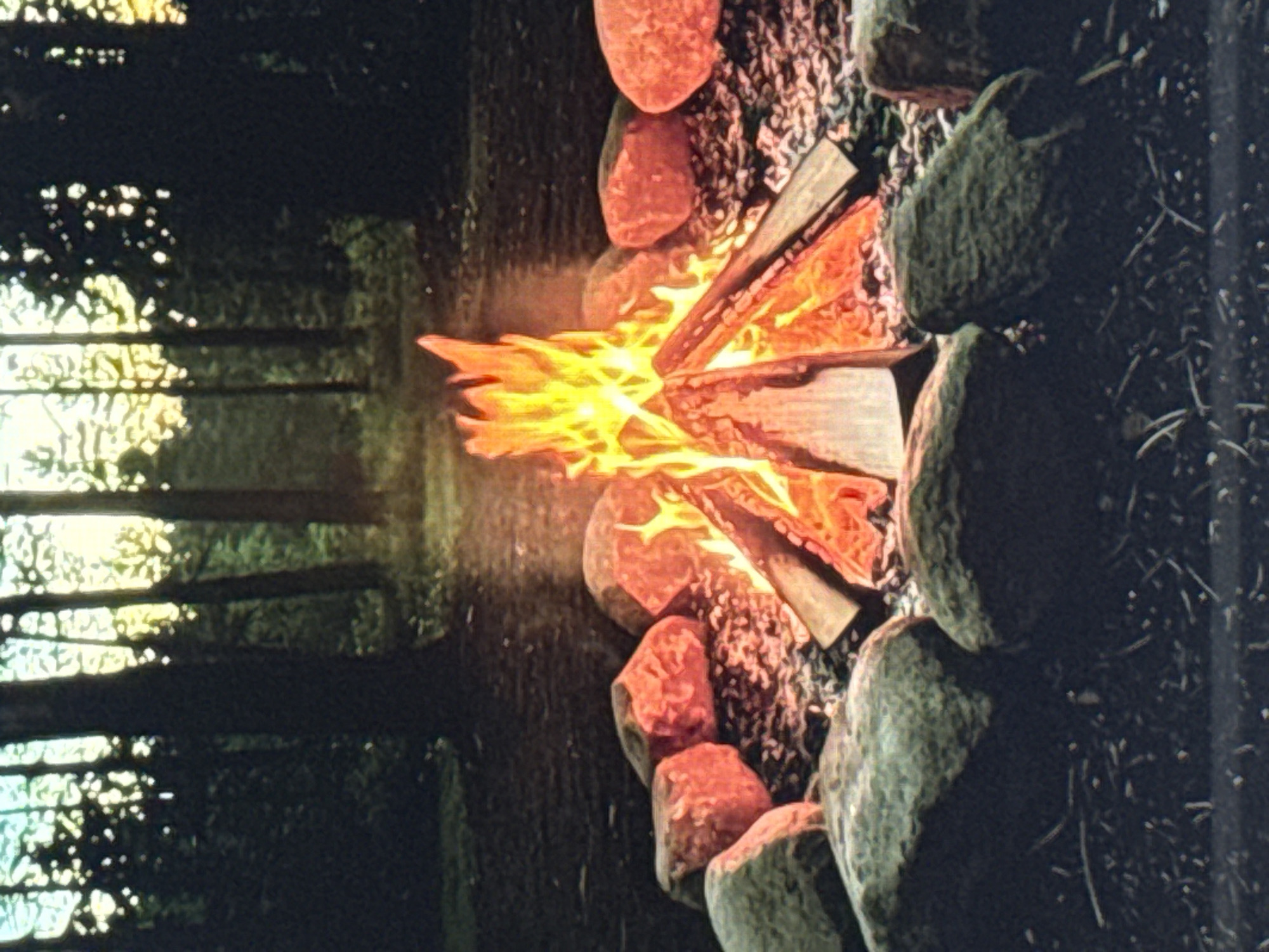 A campfire burns brightly in a forest clearing at dusk. The fire is surrounded by large rocks, some of which are reddish in color, while others are grey. In the background, tall, dark trees stand silhouetted against a hazy, yellow-orange sky, suggesting either sunset or sunrise. The ground is covered in dark soil and scattered with small twigs and debris. There are no people visible in the image. The scene is quiet and still, evoking a sense of wilderness and solitude.
