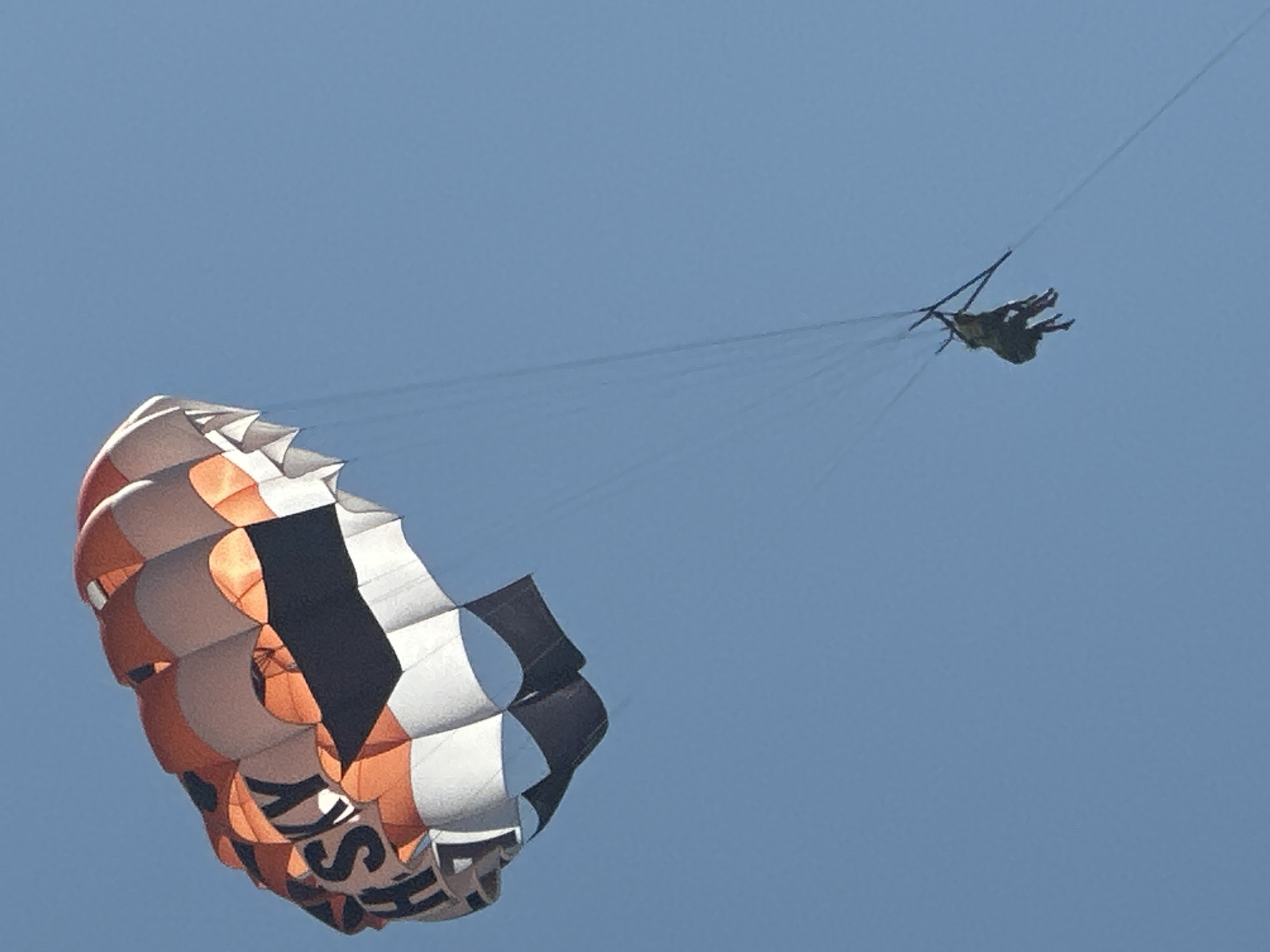 The image shows a parasailer with two people suspended beneath a large, colorful parachute. The parachute is predominantly orange and white with black accents and appears to have the word "SKY" printed on it. The parasailer is airborne against a clear blue sky, with no visible land features or city elements of Boreti, Montenegro. The scene suggests an activity of recreational parasailing.