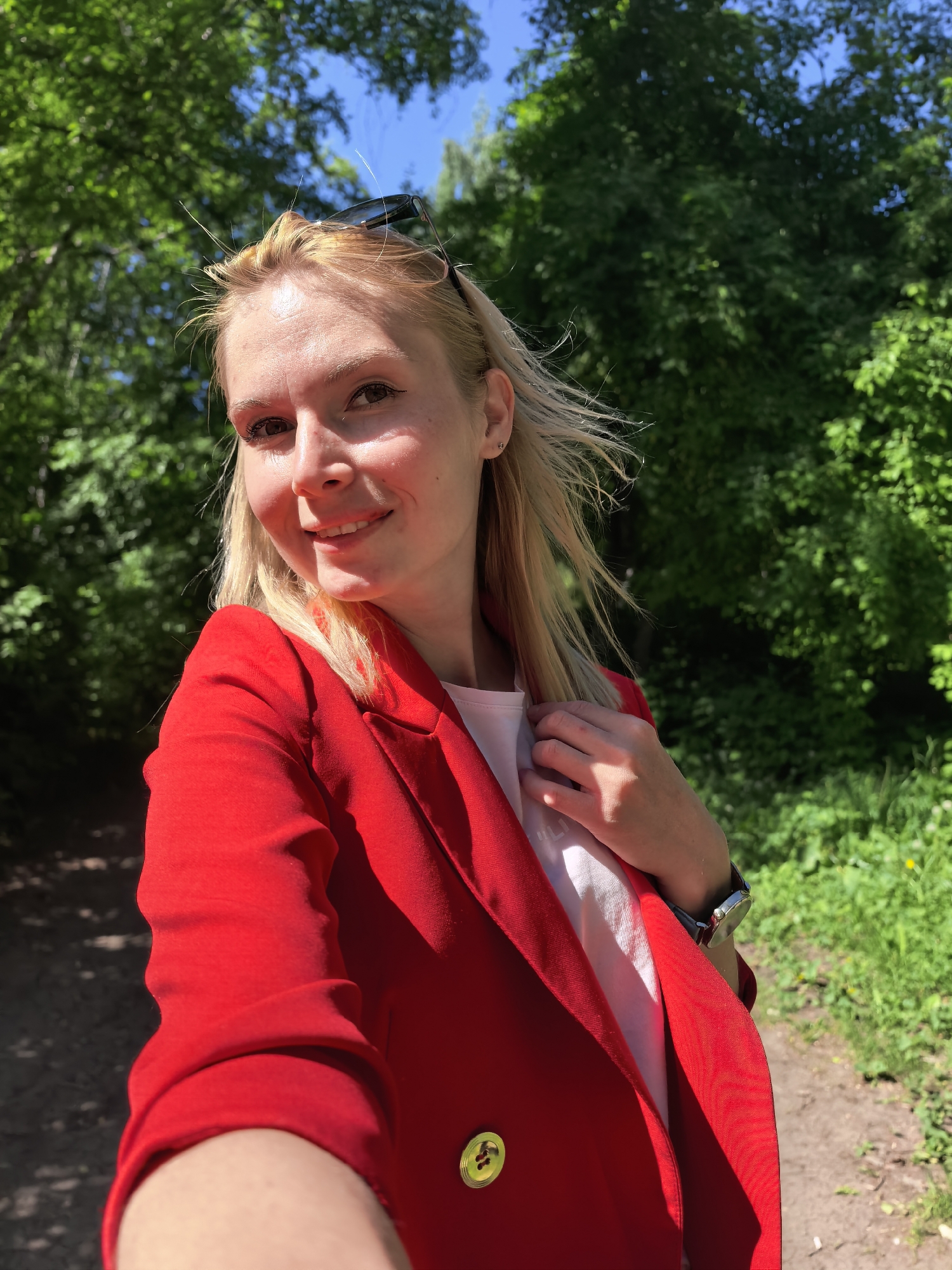 A woman takes a selfie outdoors. She is blonde, wearing a red blazer over a white top. Sunglasses are perched on her head. She is smiling slightly. She has a watch on her wrist. The background is a green forest with a trail visible on the ground. The location is Sterlitamak, Russia.