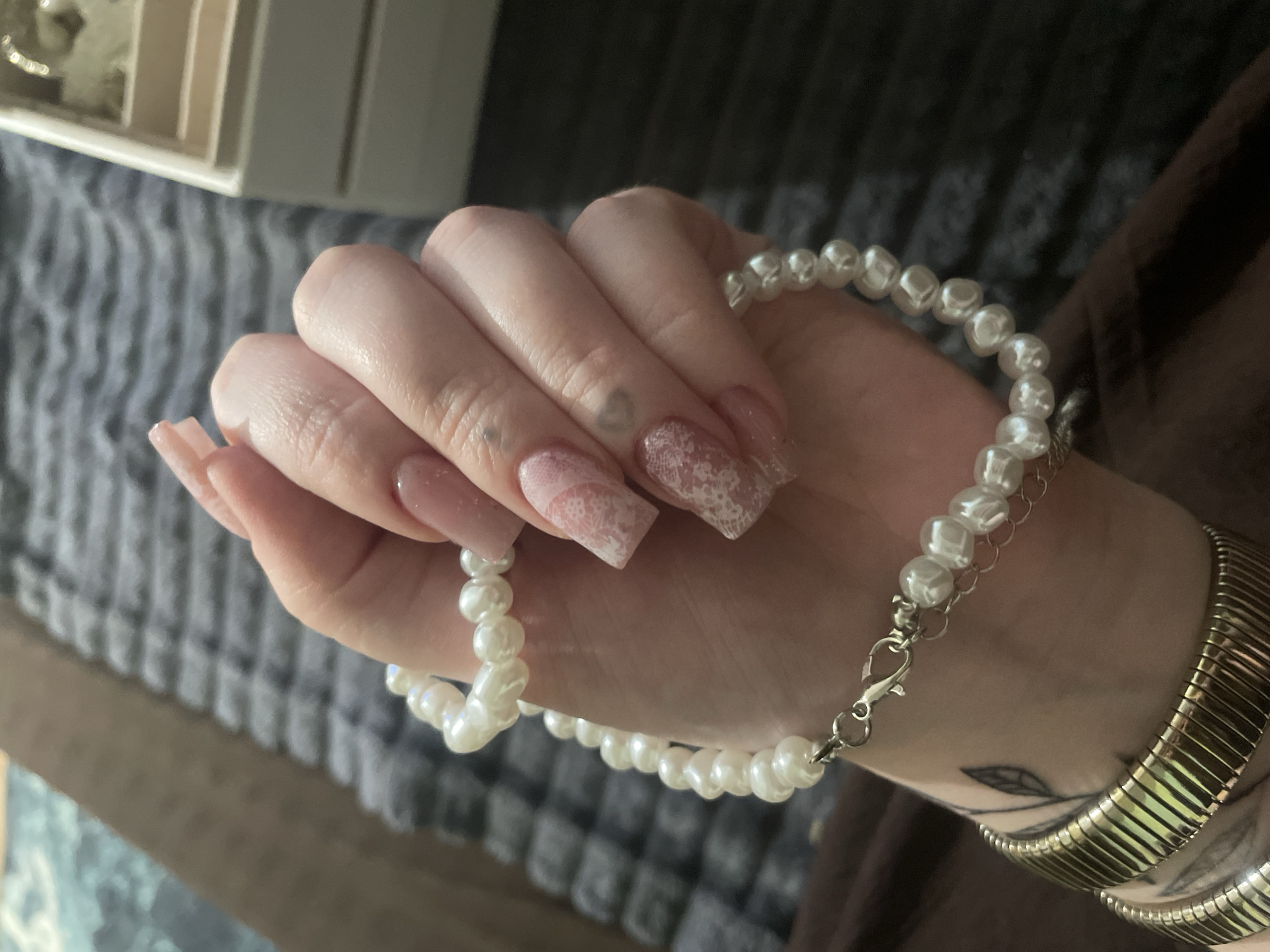 A close-up, high-angle shot shows a left hand with a pearl bracelet and a gold cuff bracelet. The hand has acrylic nails with a pinkish hue and a lace-like design on two nails. The background is a textured gray surface. There is no discernible location in the city of Линёвка, Russia.