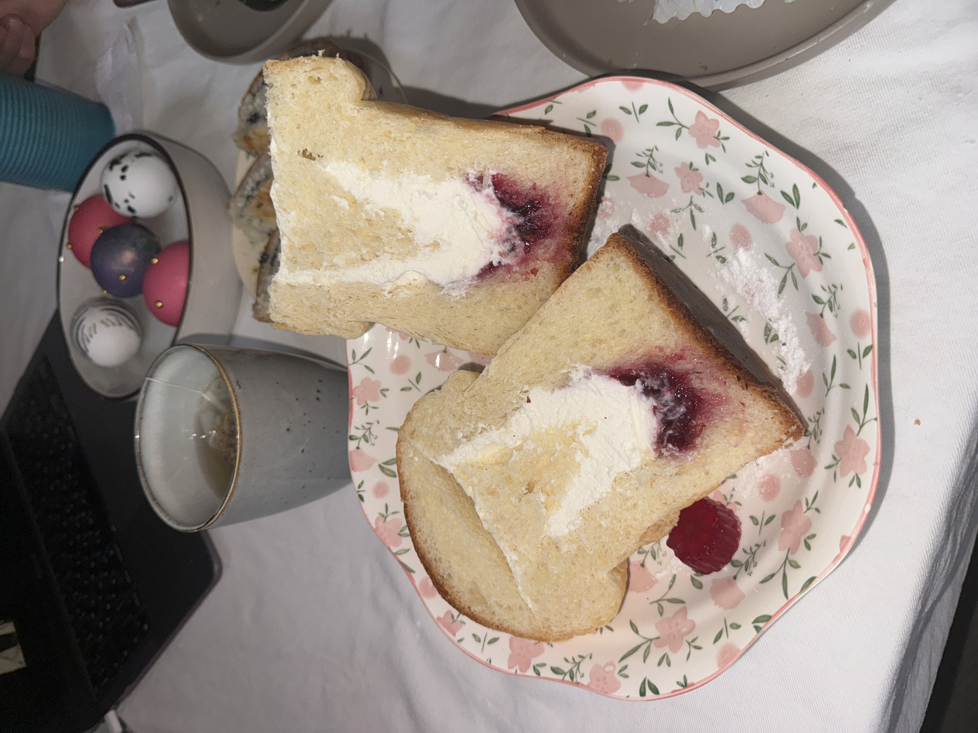 The image shows a tabletop scene with a plate of cream and jam sandwiches. Two triangle-shaped sandwiches are on a white plate with a pink floral pattern. The sandwiches are made of white bread, topped with white cream and a layer of red jam. A single raspberry sits on the plate near one of the sandwiches. A grey ceramic mug with a beverage is to the left of the plate, alongside a small bowl containing multiple colored eggs. Parts of a laptop are visible on the left of the frame. The background includes a white tablecloth. The scene appears to be indoors, on a table prepared for a meal. There is no indication of the location being in Krasnoyarsk, Russia or any specific city. No people, interactions, or actions are visible in the frame.