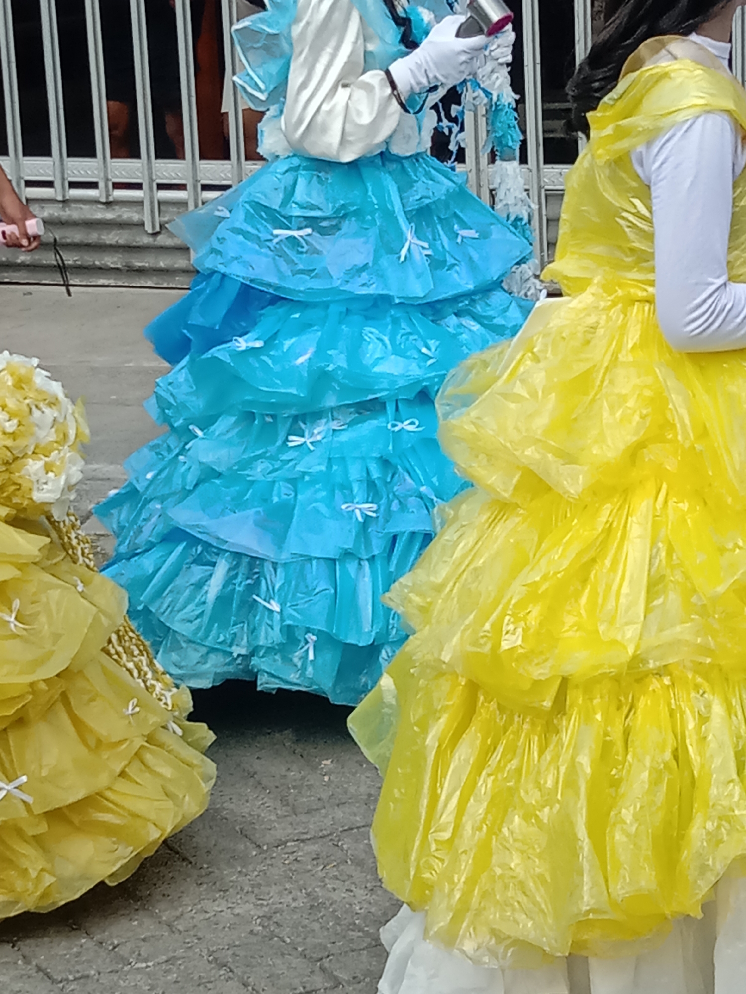 The image captures a close-up view of individuals dressed in elaborate costumes made of plastic bags. The costumes are designed with multiple layers of ruffled fabric, creating voluminous skirts. One costume is predominantly blue, featuring a layered skirt adorned with small white bows. Another costume is bright yellow, also with layered ruffles, and appears to be a similar design. A portion of a third costume, also yellow, is visible on the left side of the frame. The individuals are likely participating in a parade or festival, given the festive nature of the costumes. The background shows a metal fence and a walkway made of concrete pavers, suggesting an outdoor urban or public space. The scene is illuminated by natural light. While the image is from Wirosari, Indonesia, this specific location within the city is not discernible from the visual information provided.
