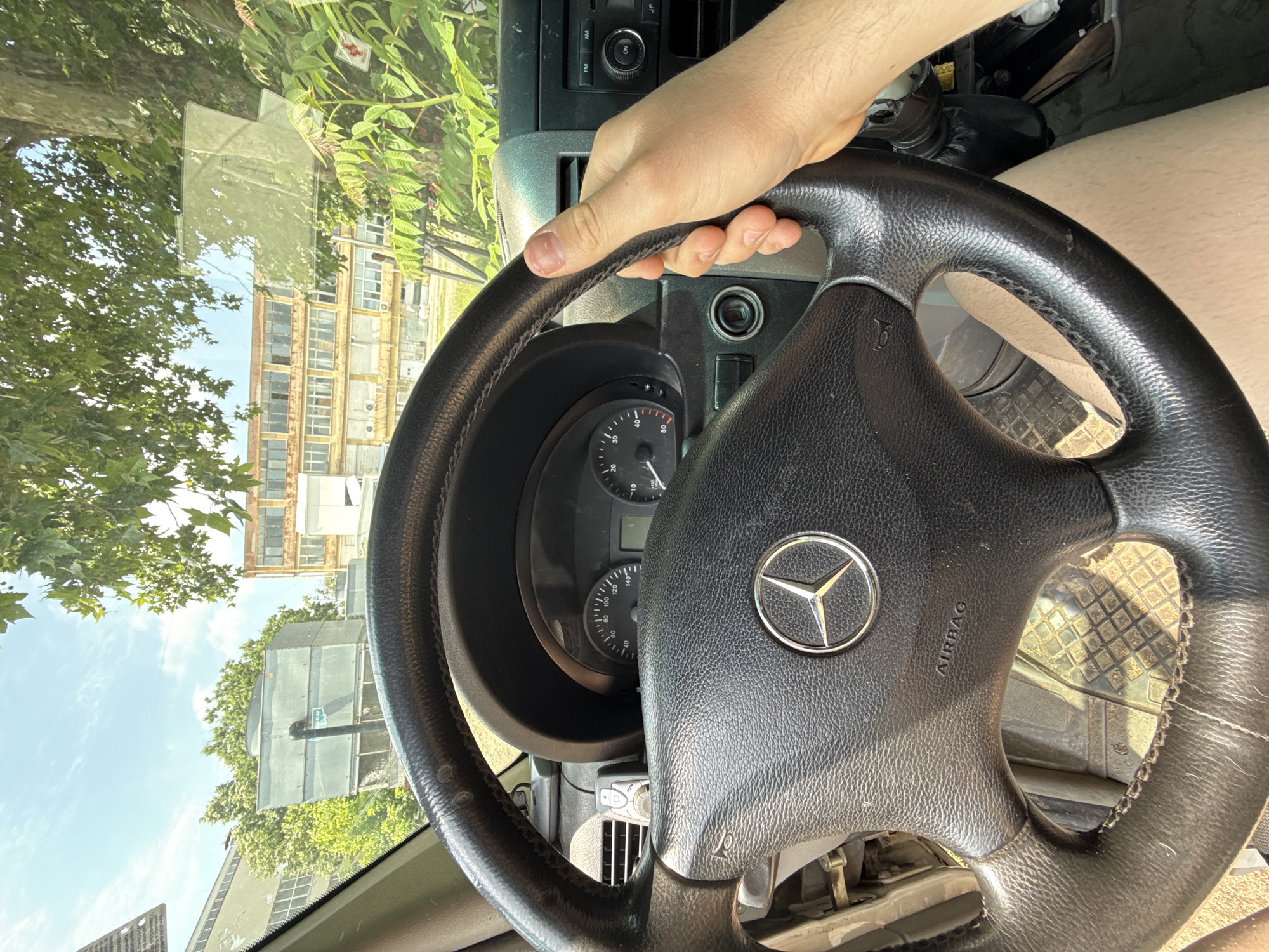 The image captures a first-person perspective from inside a Mercedes-Benz vehicle, likely a van or light commercial truck, looking out through the windshield. A person's right hand firmly grasps the steering wheel, which features a prominent Mercedes-Benz logo in its center and the word "AIRBAG" embossed on the spoke. The steering wheel itself shows signs of wear, particularly on the leather-wrapped rim.
The dashboard and instrument cluster are visible, showing a speedometer with markings up to at least 140 km/h and other gauges. To the right of the steering wheel, a radio unit with "FM AM" and "ON" buttons is present, and below it, a small electronic device is attached to the dashboard. The footwell area, with a checkered floor mat, is also partially visible.
Outside the vehicle, a bright sunny day is evident with a clear blue sky. Lush green trees, possibly plane trees given their broad leaves, frame the view. Beyond the trees, several multi-story buildings with numerous windows, appearing somewhat aged or industrial, dominate the background. One building has a distinct reddish-brown facade. A traffic light or sign pole is also visible amidst the foliage. A small white square sticker with a red cross and some illegible text is affixed to the windshield, high on the right side. Another clear sticker with illegible text is visible lower down on the windshield. The scene depicts an urban or semi-urban environment, consistent with Stara Zagora, Bulgaria.