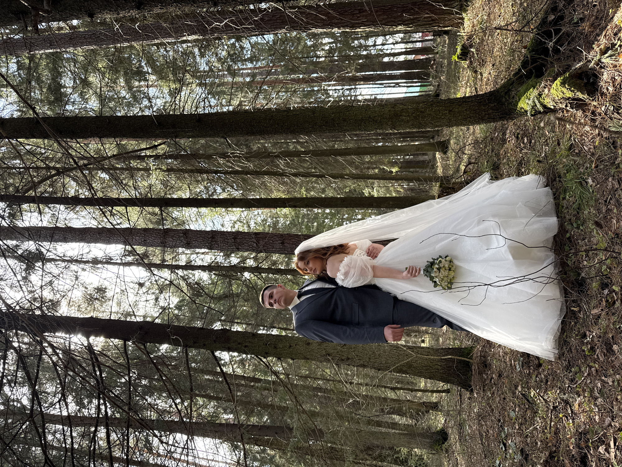 A full-length, vertically oriented photograph captures a bride and groom posing in a sun-dappled coniferous forest in Chaikovsky, Russia. The image is taken from a slightly low angle, looking up towards the towering trees.
The bride, positioned on the right, wears an elaborate white wedding gown with a voluminous skirt, sheer sleeves, and a long veil cascading behind her. She has long, light brown hair and holds a bouquet of white and cream flowers with green foliage in her left hand. She is leaning her head gently on the groom's left shoulder, looking towards the camera with a serene expression.
The groom, standing to the left of the bride, wears a dark suit, a white shirt, and a dark tie. He has short, dark hair and looks directly at the camera with a serious yet composed demeanor. His right hand is resting by his side.
The couple is surrounded by a dense forest of tall, slender pine or fir trees, their trunks dark and textured. Sunlight filters through the canopy, creating bright patches and deep shadows on the forest floor, which is covered with dry leaves, pine needles, and scattered patches of moss. The light illuminates the bride's white gown, making it stand out against the earthy tones of the environment. No text is visible in the image. The scene suggests a romantic wedding photoshoot taking place outdoors, likely during the day.