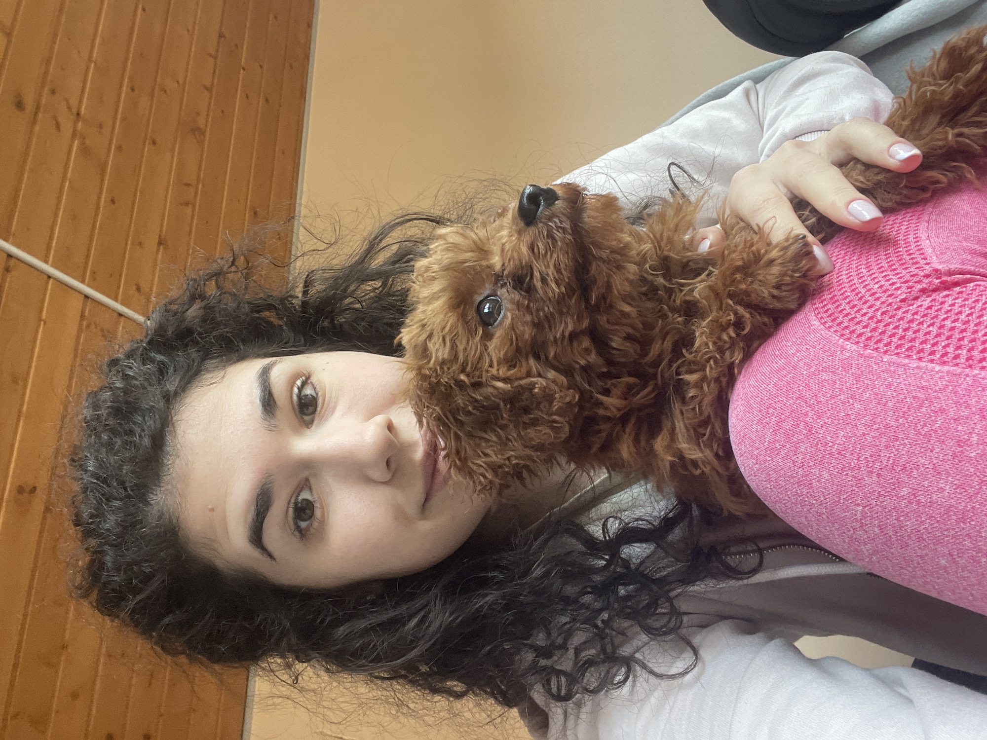 A young woman with dark, curly hair is holding a brown poodle close to her face. She is looking directly at the camera with a slight smile. She is wearing a white hoodie and pink leggings. The background includes a wooden wall and a light peach-colored wall. The location is not determinable from the image.