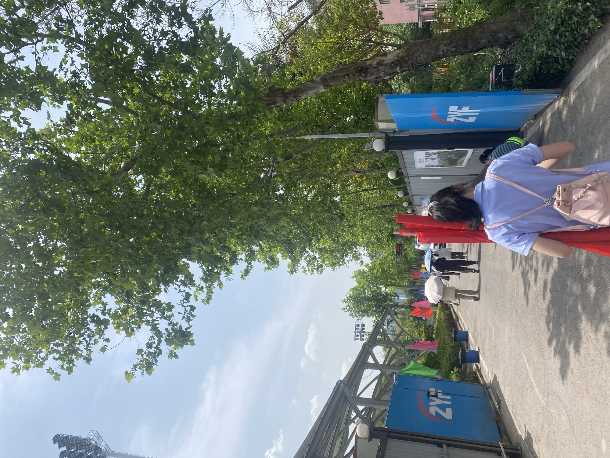 The image shows an outdoor scene in the Chongqing High-tech Zone, China. A pedestrian walkway, lined with trees forming a green canopy, is visible under a partly cloudy sky. There are blue banners with white lettering on the right side of the walkway, and red banners further down. People are walking along the walkway, some holding umbrellas. A woman in a light purple outfit with a light pink backpack stands along the side. In the background, there is a building and lighting.
