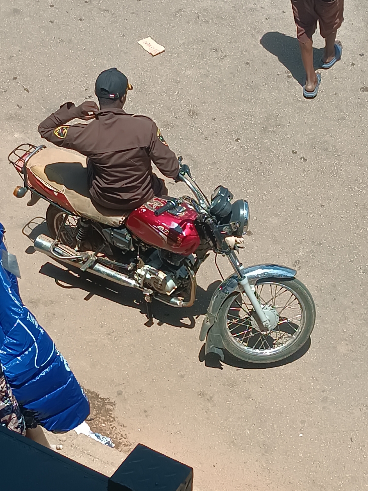 The image depicts a man wearing a brown uniform and a black cap, seated on a red and chrome motorcycle. The uniform has patches on the sleeves. The motorcycle is parked on a paved surface, possibly a street. Another person's legs and feet, wearing shorts and sandals, are visible in the upper right corner of the image. A large, blue, puffy object is visible in the lower left. There is also a small, light-colored rectangular object on the pavement. This location is Bukuru, Nigeria.