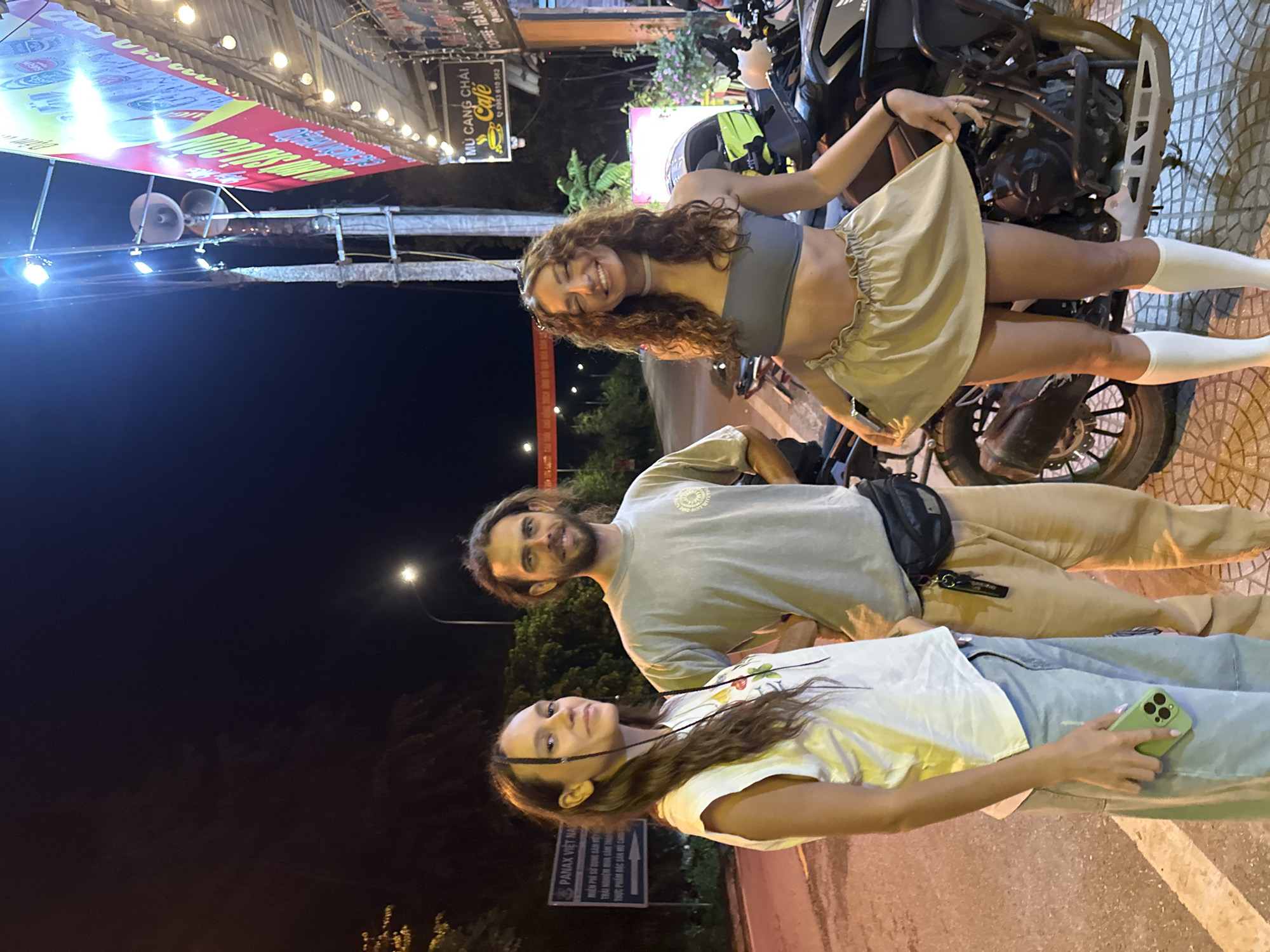 This image depicts three individuals posing for a photograph at night on a paved street or sidewalk, likely in Mù Cang Chải, Vietnam. The scene is brightly lit by overhead lights and storefronts, contrasting with the dark night sky in the background.
In the foreground, a young woman with long, dark hair, some of which is braided, stands wearing a white t-shirt and light blue jeans. She holds a light green smartphone in her right hand and looks towards the camera with a neutral expression. Beside her, a man with brown, shoulder-length hair and a beard wears a light grey t-shirt and tan pants, with a black fanny pack or small bag slung across his body. He offers a slight smile to the camera. Behind them, a second young woman with long, curly brown hair leans against a dark-colored motorcycle. She is dressed in a grey crop top, a light beige ruffled mini-skirt which she slightly lifts with one hand, and white knee-high socks. She smiles broadly towards the camera.
The time of day is clearly night, indicated by the dark sky and the reliance on artificial light sources. The atmosphere appears relaxed and friendly, with all three subjects looking directly at the viewer.
Several signs are visible in the illuminated background. A prominent banner displays "MÙ CANG CHẢI Cafe" with a coffee cup logo and the phone number "0983 610 562." Other partial Vietnamese text, possibly "COFFEE - TRÀ SỮA," is also present on this banner. Further down, another sign clearly reads "PANAX VIỆT NAM." The setting suggests a casual evening stop in a commercial area.