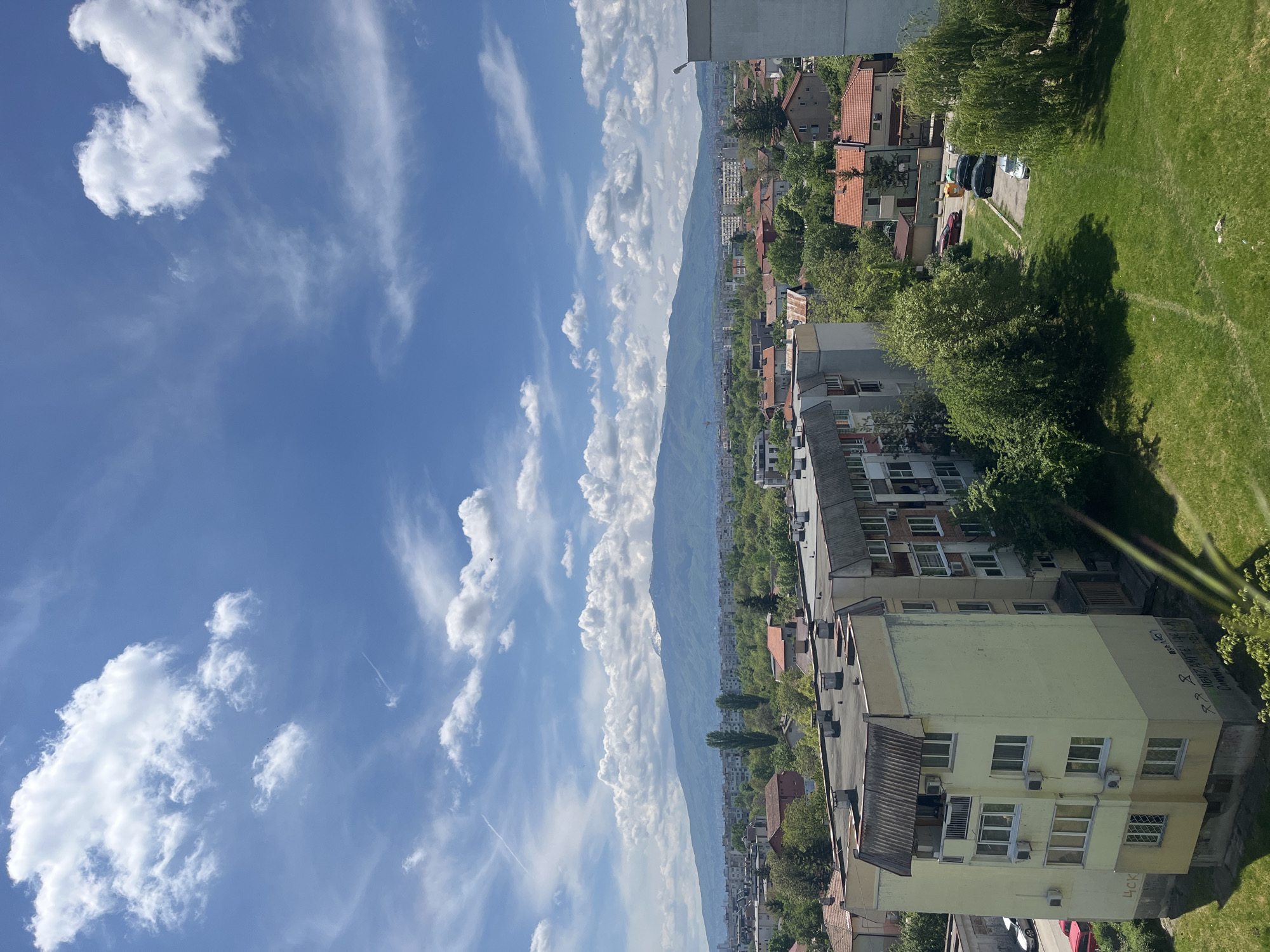 The image presents a cityscape view in Sofia, Bulgaria, facing towards the mountains. The foreground is dominated by multi-story buildings with apartments, some showing signs of age with visible air conditioning units and a green/yellowish facade. There are sections of green lawns and trees bordering the building edges. The middle ground shows a series of residential buildings interspersed with trees, leading to the backdrop of a mountain range under a bright, blue sky with scattered clouds. The sky takes up the upper half of the image, displaying a bright, clear blue with some white cirrus and cumulus clouds.