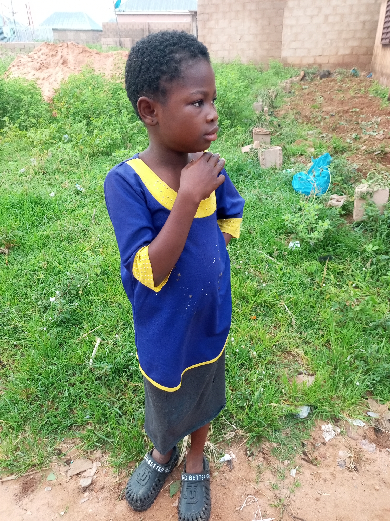 A young Black child, likely under ten years old, stands facing right in an outdoor environment. The child has short, dark, curly hair and dark skin. They are wearing a blue shirt with yellow trim around the collar and sleeves, and a dark grey skirt. The child's right hand is raised and touching their chin, and their gaze is directed off-camera to the right. The child is wearing black slip-on sandals with "GO BETTER" printed on the side in white text.
The setting appears to be a somewhat unkempt, possibly semi-urban or rural area. The ground is a mix of dirt and patchy green grass. In the background, there is a pile of reddish-brown dirt or sand to the left, and a brick building under construction or in disrepair to the right. Some scattered building materials, including concrete blocks and a crumpled blue plastic bag, are visible in the mid-ground. The overall impression is of an undeveloped or developing area. The location is identified as Tempure, Nigeria.