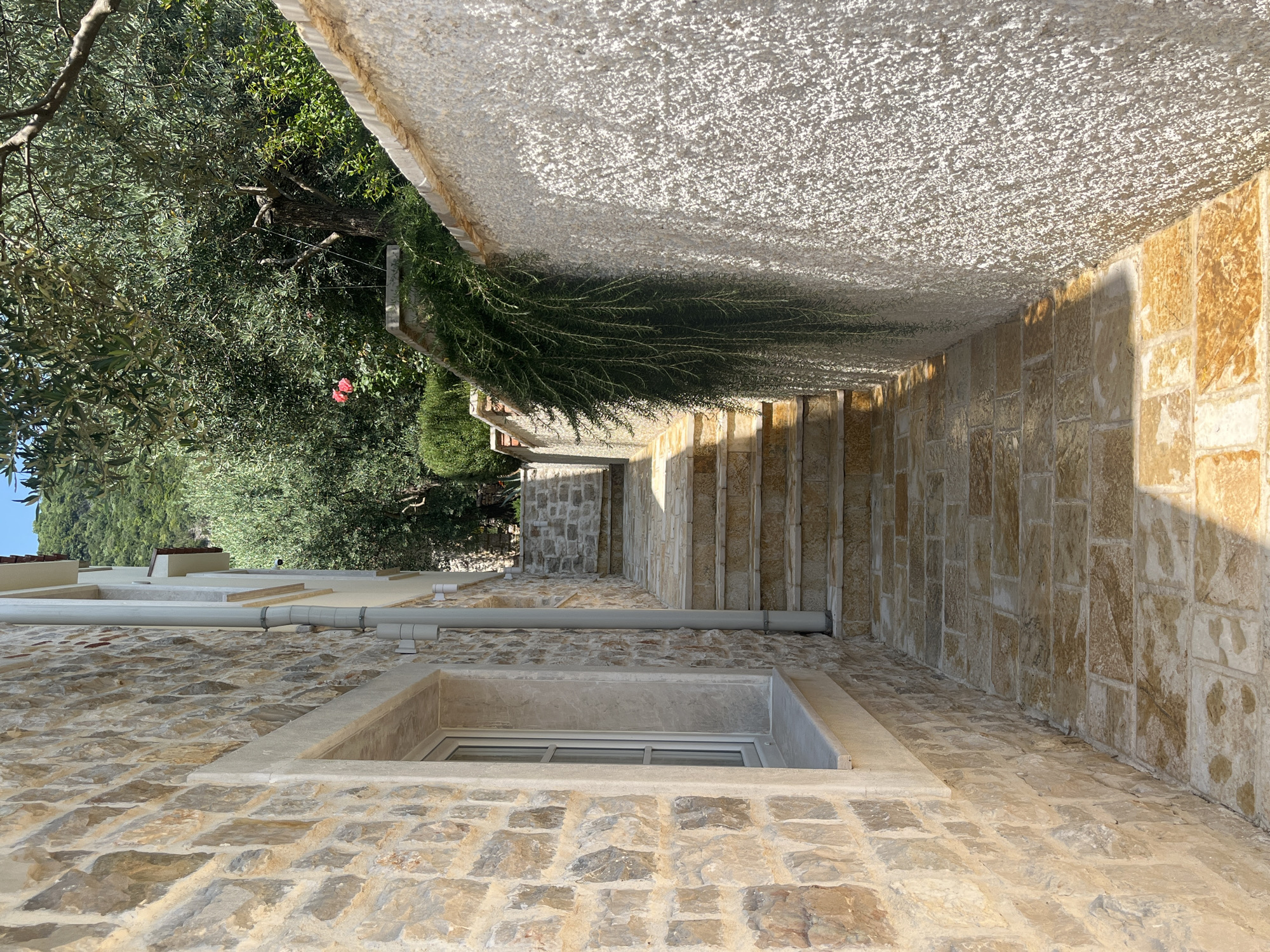 The image shows an outdoor scene with a stone-built structure. The perspective is from a low angle, looking up towards a stone staircase and an overhead covered walkway or ceiling made of white, rough-textured material. The walls are constructed from large, irregular light brown and beige stones. A small window with a white frame is visible in the stone wall in the foreground. Above the window, grey pipes run along the wall.
To the left, a lush green tree with dark foliage and branches extends into the frame, with a hint of blue sky visible. Two small red flowers are nestled within the foliage. Further back, more greenery and parts of a building with a tiled roof can be seen. The stone staircase, made of the same rough, light-colored stones as the walls, leads upwards into the distance. The walkway above the stairs is illuminated by sunlight, casting shadows on the staircase.
The scene appears to be in a Mediterranean or similar climate, given the stone architecture, olive tree, and bright sunlight. The time of day is likely morning or afternoon, as indicated by the strong shadows. The overall impression is one of a charming, rustic outdoor space. There is no visible text in the image. The location is Kumbor, Montenegro.