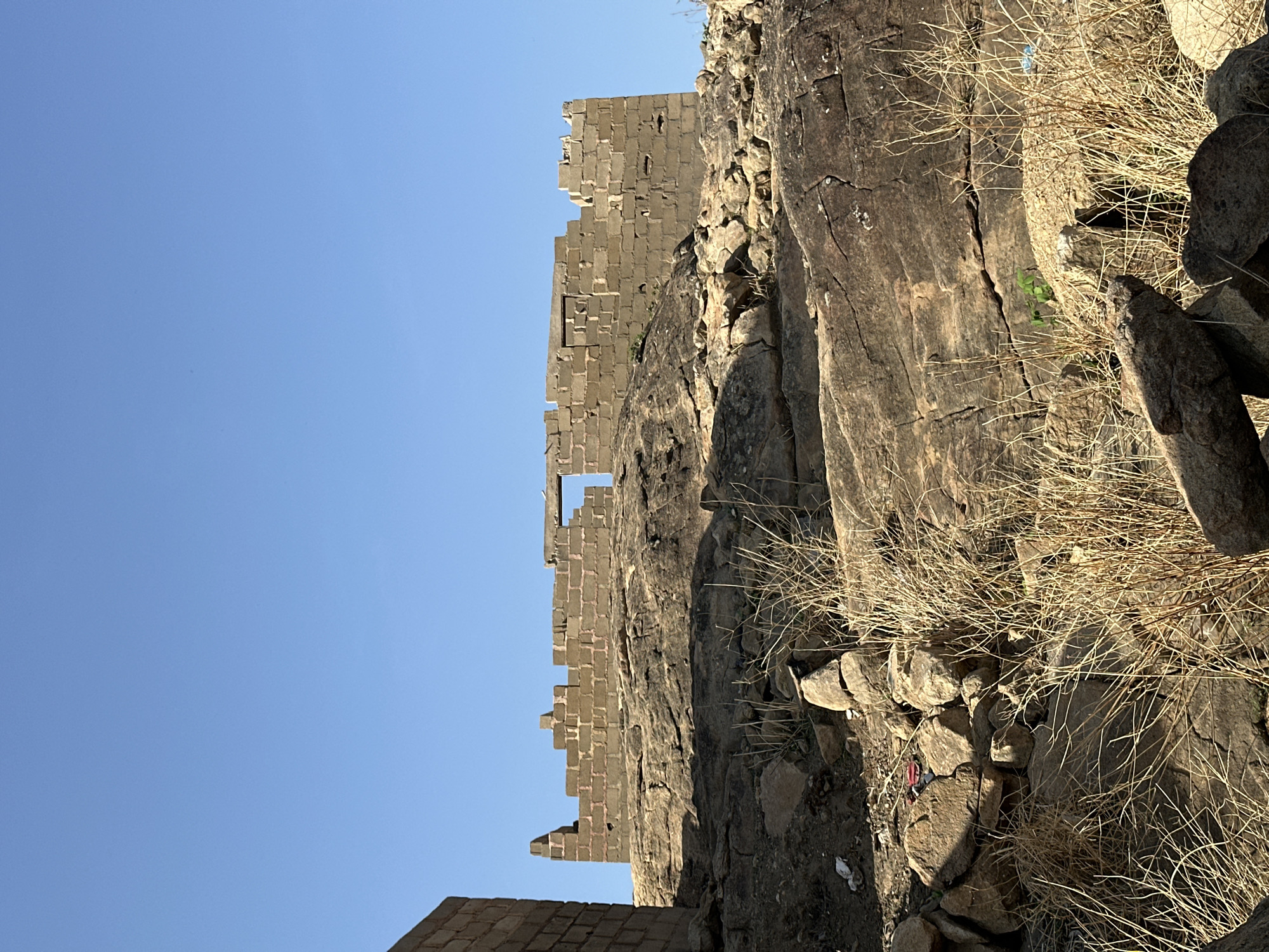 The image depicts a section of a ruined stone building with a clear blue sky in the background. The stone structure is tall and appears to be part of a wall or fortification, possibly an old castle. It is made of stacked, rectangular stone blocks. The structure is situated on or next to a rocky hillside, with rough, weathered rock formations visible. Sparse dry vegetation and small rocks are scattered on the terrain around the base of the structure. The scene is likely an arid or semi-arid environment. Given the location in Jos, Nigeria, it is likely a view of the Shere Hills, where there are many rocky outcrops. There are no people visible in the image.