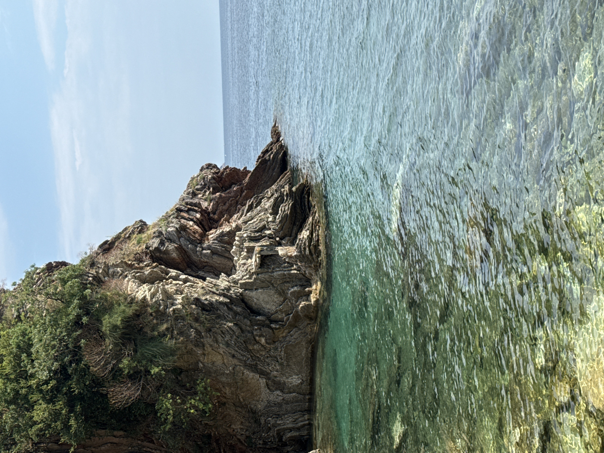 The image depicts a rocky coastline with clear turquoise water. The scene is oriented vertically, with the sea occupying the right half and a rocky cliff face covered in green foliage on the left. The water is shallow and clear, revealing the seabed or submerged rocks beneath its surface. Sunlight creates shimmering patterns on the water, indicating a bright day.
The cliff is rugged and layered, with a mix of stone and sparse vegetation clinging to its steep slopes. The foliage is dense in some areas, appearing as dark green shrubs and trees. The overall impression is one of a natural, undeveloped coastal landscape.
The sky above is a pale blue with a few wispy clouds, suggesting fair weather. The time of day appears to be daytime, likely late morning or early afternoon, given the bright lighting. There are no people or man-made objects visible in the frame. The location context of Boreti, Montenegro, aligns with the visual cues of a Mediterranean or Adriatic coastline, known for its rocky shores and clear waters. The image evokes a sense of tranquility and natural beauty. There is no visible text in the image.