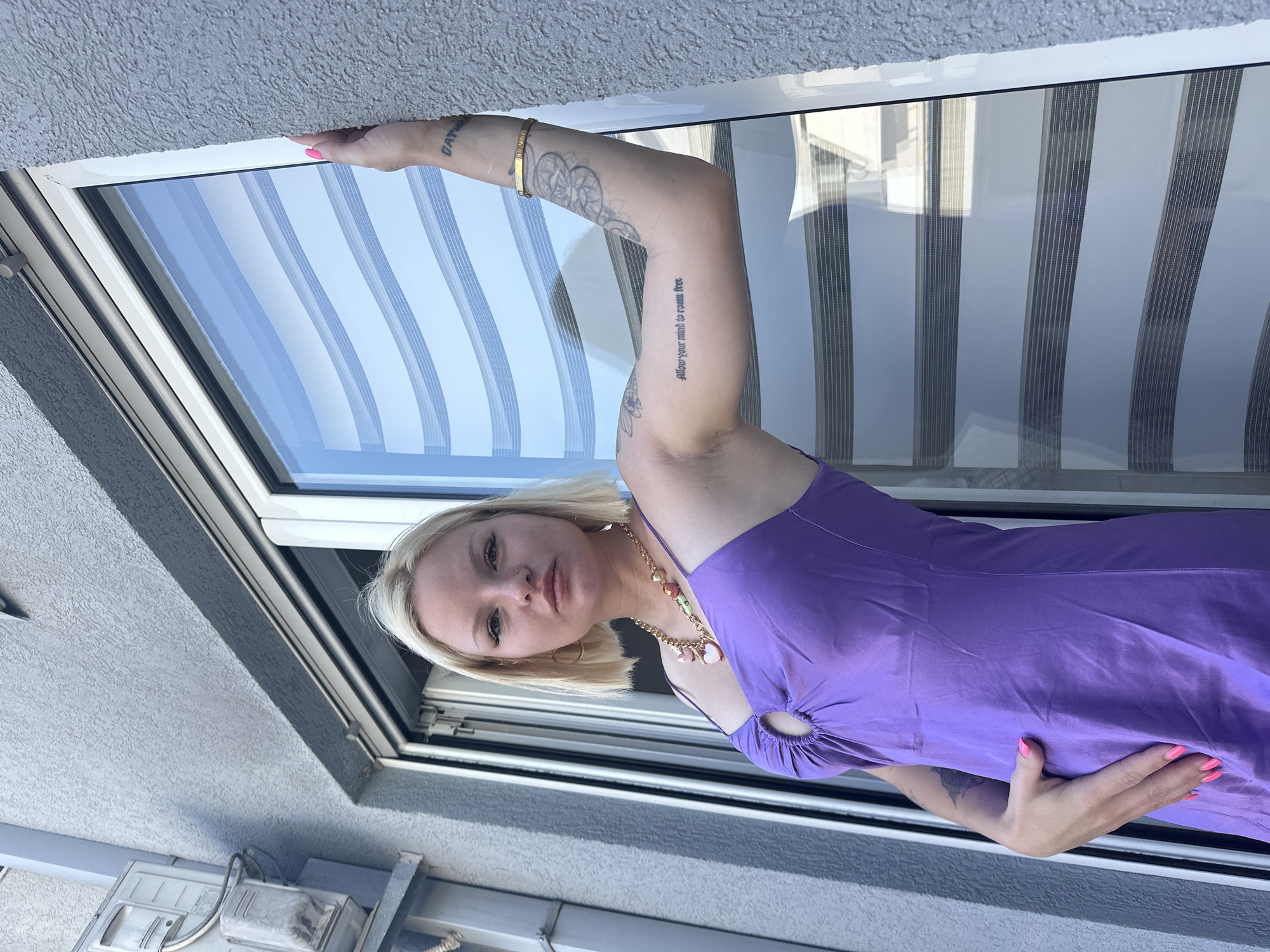 In Budva, Montenegro, a young blonde woman with a bob haircut and minimal makeup poses by a window. She is wearing a purple spaghetti strap dress with a cut-out detail at the bust and a gold necklace. Her right arm is extended, with a tattoo visible on her forearm and a gold bracelet on her wrist. Her left hand rests on her stomach. The background consists of a gray textured wall and a window with horizontal blinds. An air conditioning unit is visible on the wall to the left.