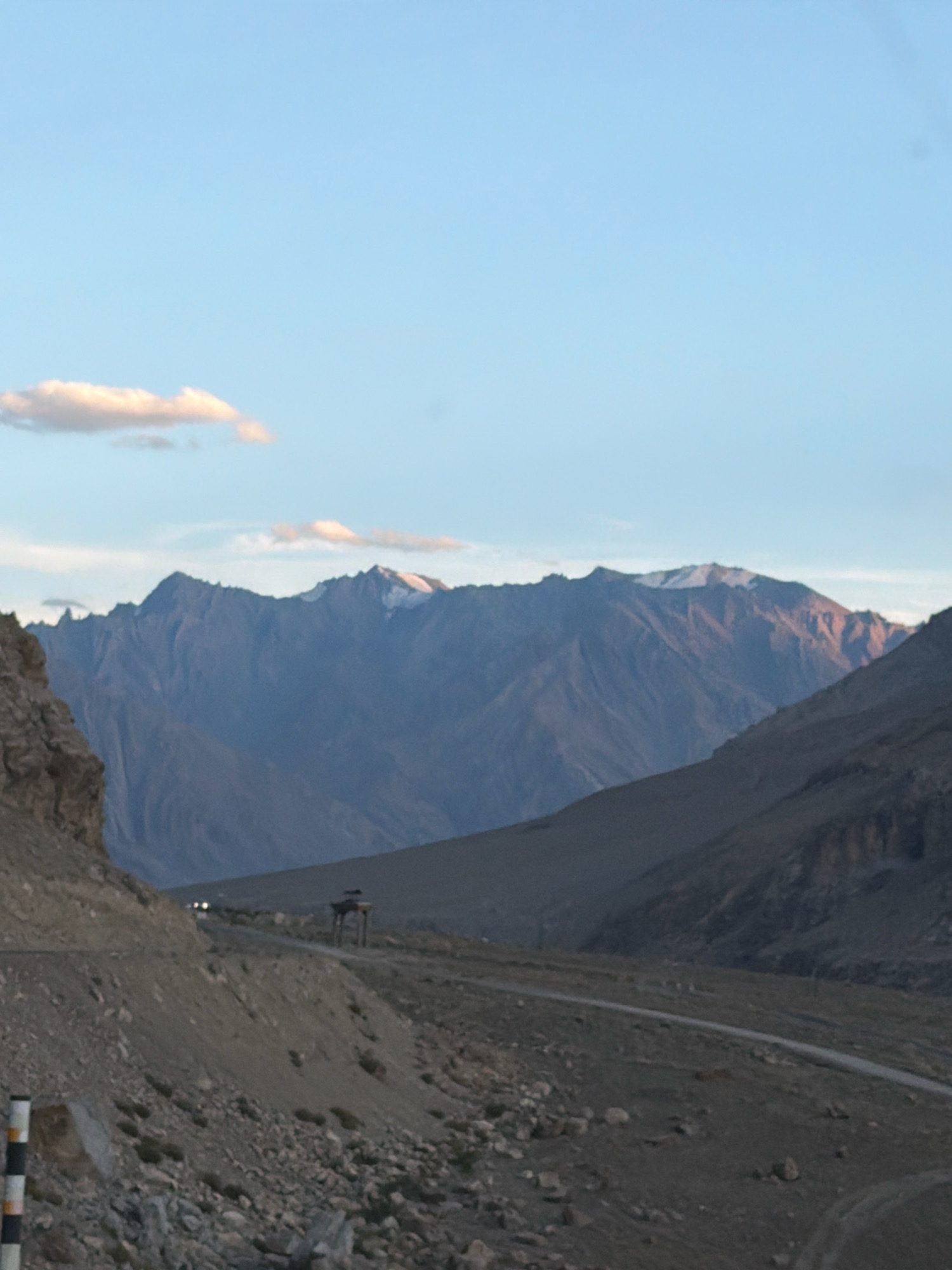 The image shows a mountainous landscape in the city of Pidmo, India. The foreground features a rocky, barren slope with a paved road winding through it. To the left, a post with black and white stripes is visible. In the middle ground, a dark structure, possibly a shelter or viewpoint, is situated on a hillside. Further in the distance, a road is visible with two small vehicles. The background is dominated by large, rugged mountains with some snow-capped peaks. The sky is clear with a few wispy clouds. The overall scene depicts a remote, high-altitude environment.
