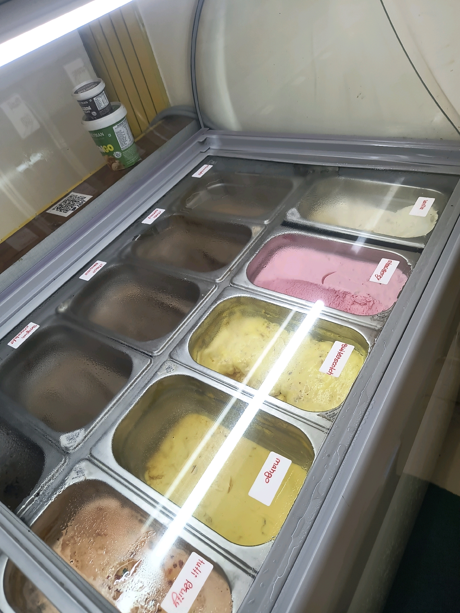 The image displays an interior view of a commercial ice cream freezer, likely located in a shop or restaurant in Hyderabad, India. The freezer is filled with various tubs of ice cream, each labeled with its flavor. Visible labels include "Mango," "Choco Brownie," "Belgium Chocolate," "Strawberry," "Butterscotch," and "Vanilla." Some of the ice cream tubs are covered with condensation. In the background, on a shelf above the freezer, are two cylindrical containers of what appears to be ice cream or a related product, with one container labeled "MANGO." A QR code sticker is also visible on the shelf. The lighting suggests an indoor commercial setting.