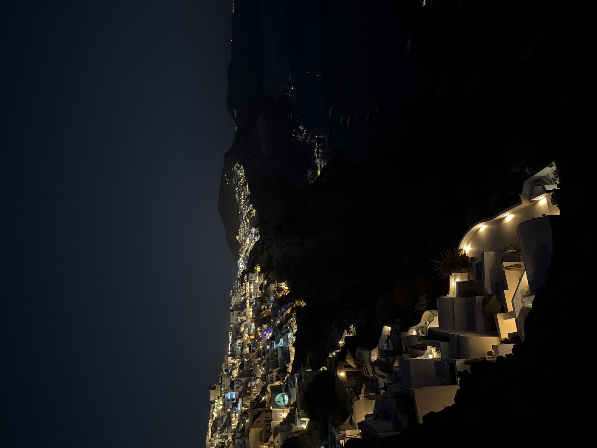 A nighttime aerial view captures the whitewashed buildings of Oia, Santorini, Greece, clinging to a steep cliffside overlooking the Aegean Sea. Lights from homes, businesses, and streetlamps illuminate the village, creating a warm glow against the dark sky. The sea below is dotted with the lights of several boats. The architecture is characteristic of Cycladic design, with cubist structures and domed roofs. The natural landscape is rugged and dramatic, with the village built into the contours of the volcanic caldera. The scene is serene and picturesque, embodying the iconic beauty of Santorini at night.
