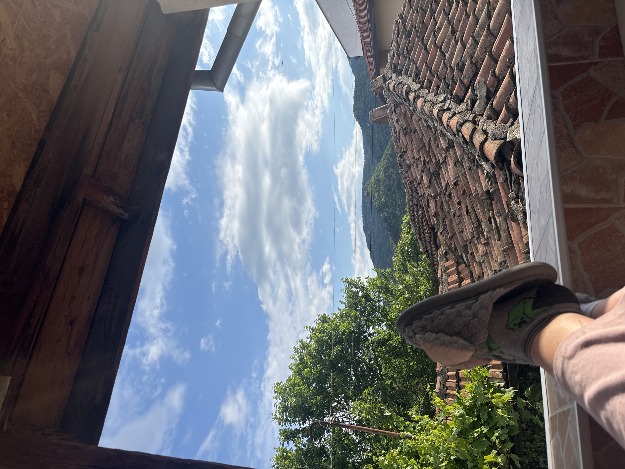 A view from a window in Asenovgrad, Bulgaria, shows a blue sky with scattered white clouds. A mountain covered in green trees is visible in the distance. The window frame is made of wood on one side, while the other side has a clay tile roof. A person wearing a pink shirt is holding a slipper with a gray and green design near the window. Green trees are visible below the roof.