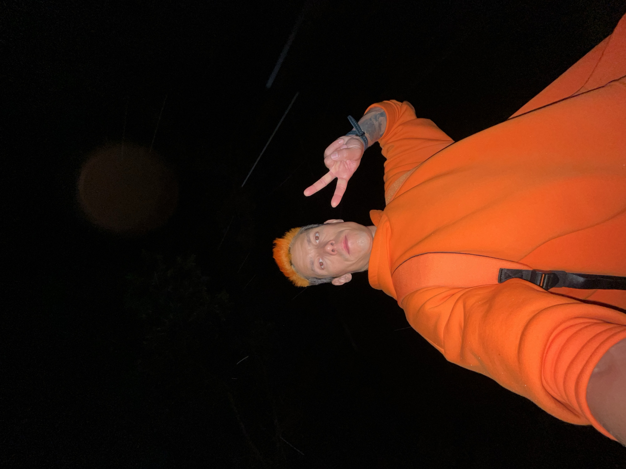 A low-angle selfie depicts a male individual with short, spiky, bright orange hair, positioned in the foreground against a dark, nocturnal background. He wears a vibrant orange hooded sweatshirt, with a dark backpack strap visible over his right shoulder. His right hand is raised, forming a peace sign (V-sign), and a dark-colored wristband or watch is present on his right wrist, above a visible forearm tattoo. His expression is neutral, and his face shows slight moisture. The background is predominantly black, indicating low light conditions, with faint, blurred streaks of light in the upper right quadrant and numerous small, bright specks scattered throughout the scene, possibly illuminated precipitation or dust. A large, diffuse, brownish-orange circular light anomaly is present in the upper center of the frame. The scene is located in Chaikovsky, Russia.