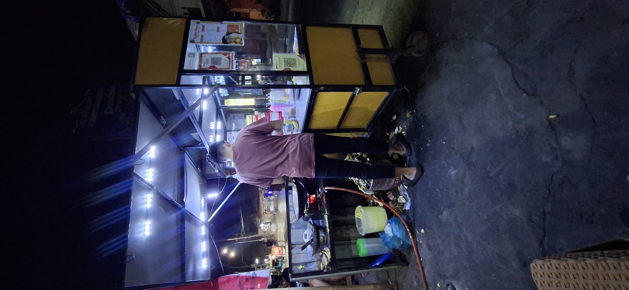 The image depicts a nighttime scene at a food stall in Klumprit, Indonesia. A person, viewed from behind, is standing and engaged in food preparation. They are wearing a maroon t-shirt and dark pants, with black flip-flops. The food stall itself is illuminated by bright LED lights from above and features a yellow and black structure. On the counter, there are various bottles, likely containing condiments, and a glass display case showcasing food items. To the left of the stall, a stove with a wok is visible, with a fire burning underneath. Several containers and bags are also present in this area, along with some discarded banana peels on the ground. The ground surface appears to be asphalt or a similar dark material, with some cracks and debris. In the background, a street with passing vehicles and streetlights suggests the stall is located in a public area.
