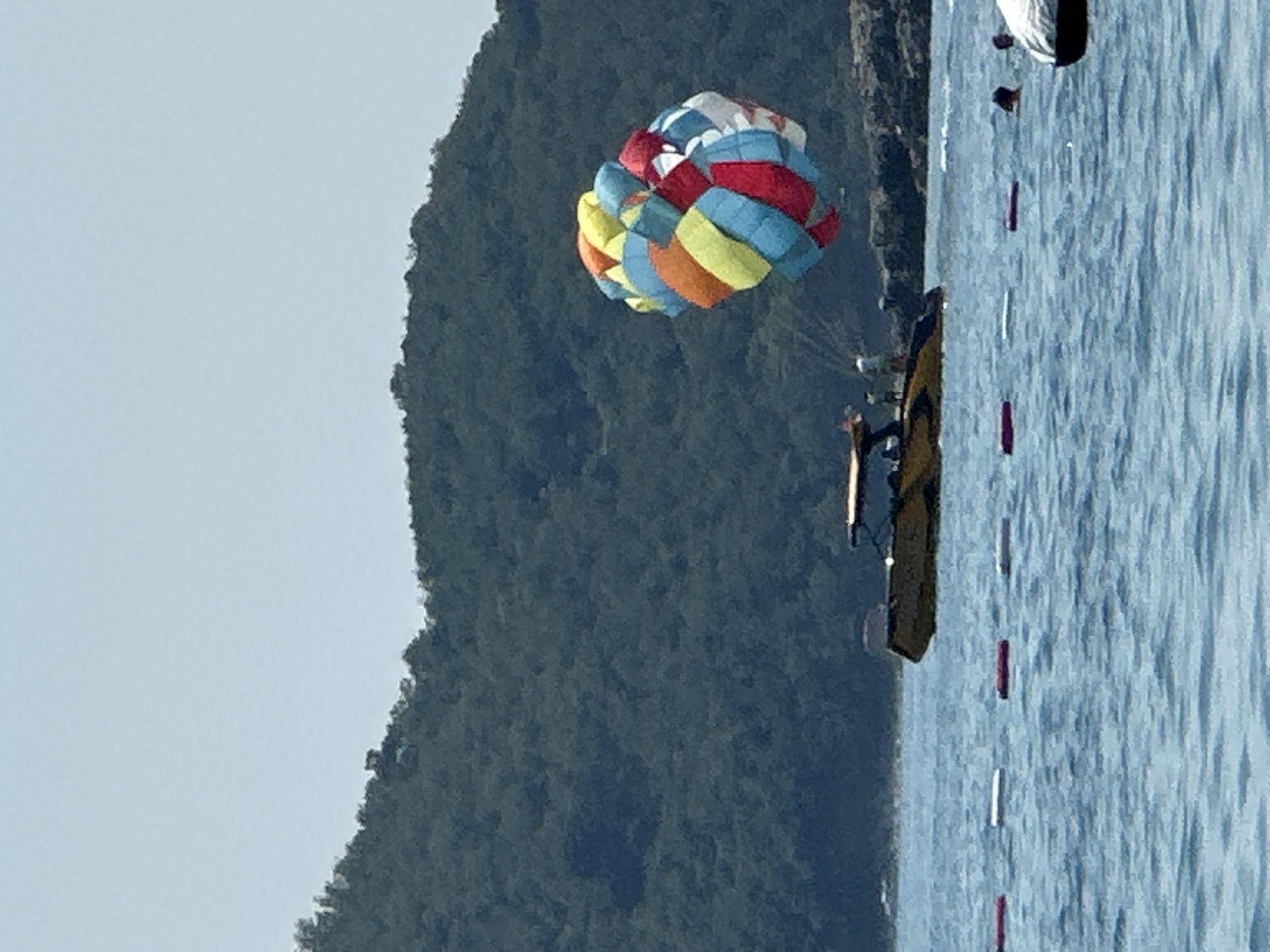 This media file, shot in Boreti, Montenegro, captures a scene of parasailing on a clear day. The main subjects are a colorful parasail billowing in the sky and a boat on the water. The parasail is a vibrant mix of red, blue, yellow, and orange panels, indicating it's in use for a recreational activity. Below, a boat, likely towing the parasailer, is visible on the calm, blue water. The background features a lush, green mountainside, characteristic of the Montenegrin coast.
The activity taking place is parasailing, a popular water sport where a person is towed behind a boat while attached to a parachute-like canopy. The image suggests a pleasant day, with bright daylight and clear skies, suitable for outdoor activities. The water appears relatively calm, with gentle ripples, and the overall impression is one of leisure and outdoor fun. The composition places the parasail as the focal point, with the boat and the scenic backdrop of the mountains providing context. There is no visible text in the image.