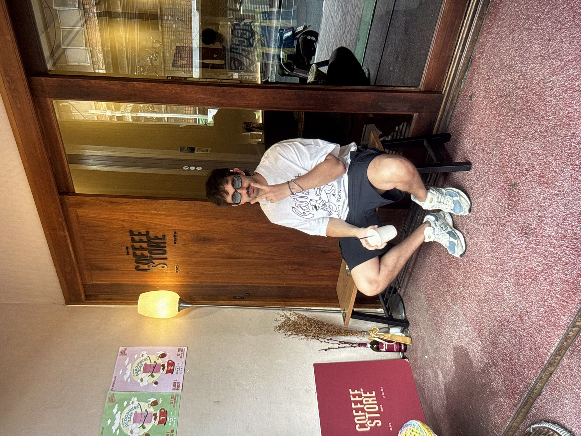 A young man with dark hair, a mustache, and sunglasses is sitting on a wooden bench outside a coffee shop. He is wearing a white t-shirt with a graphic print, dark shorts, and light-colored sneakers. He holds a white paper cup in his right hand and has his left hand raised to his chin. The coffee shop has a wooden door with "THE COFFEE STORE" written in stylized lettering and a glass sliding door. Two posters are visible on the wall to the left of the door. A floor lamp stands near the posters. The ground in front of the shop is covered with a red carpet-like material. In the reflection of the glass door, a motorcycle and a person are visible. The scene appears to be outdoors during the daytime. The location is not specified as Moscow, Russia.