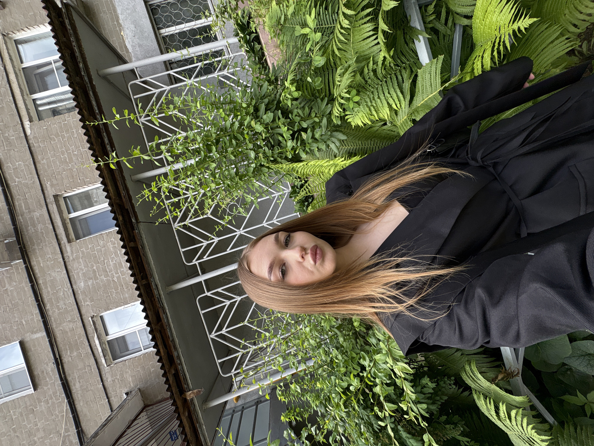 A young woman with long, blonde hair and fair skin is taking a selfie in front of an old brick building with balconies. She is wearing a black blazer with a belt. The building appears to be in a state of disrepair, with peeling paint and a rusty roof. Lush green plants, including ferns, are growing around the building. The sky is overcast. The scene is likely located in Chaikovsky, Russia, as indicated by the architectural style and the surrounding vegetation, which are common in the region.