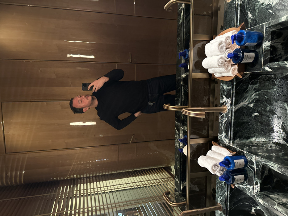 A man stands in a luxurious bathroom, taking a selfie in the mirror. He is wearing a black long-sleeved shirt and dark pants. The bathroom features brown wooden walls, marble countertops, and gold accents. There are towels and blue bottles neatly arranged on the countertop. The mirror reflects the man and the stylish interior of the bathroom.