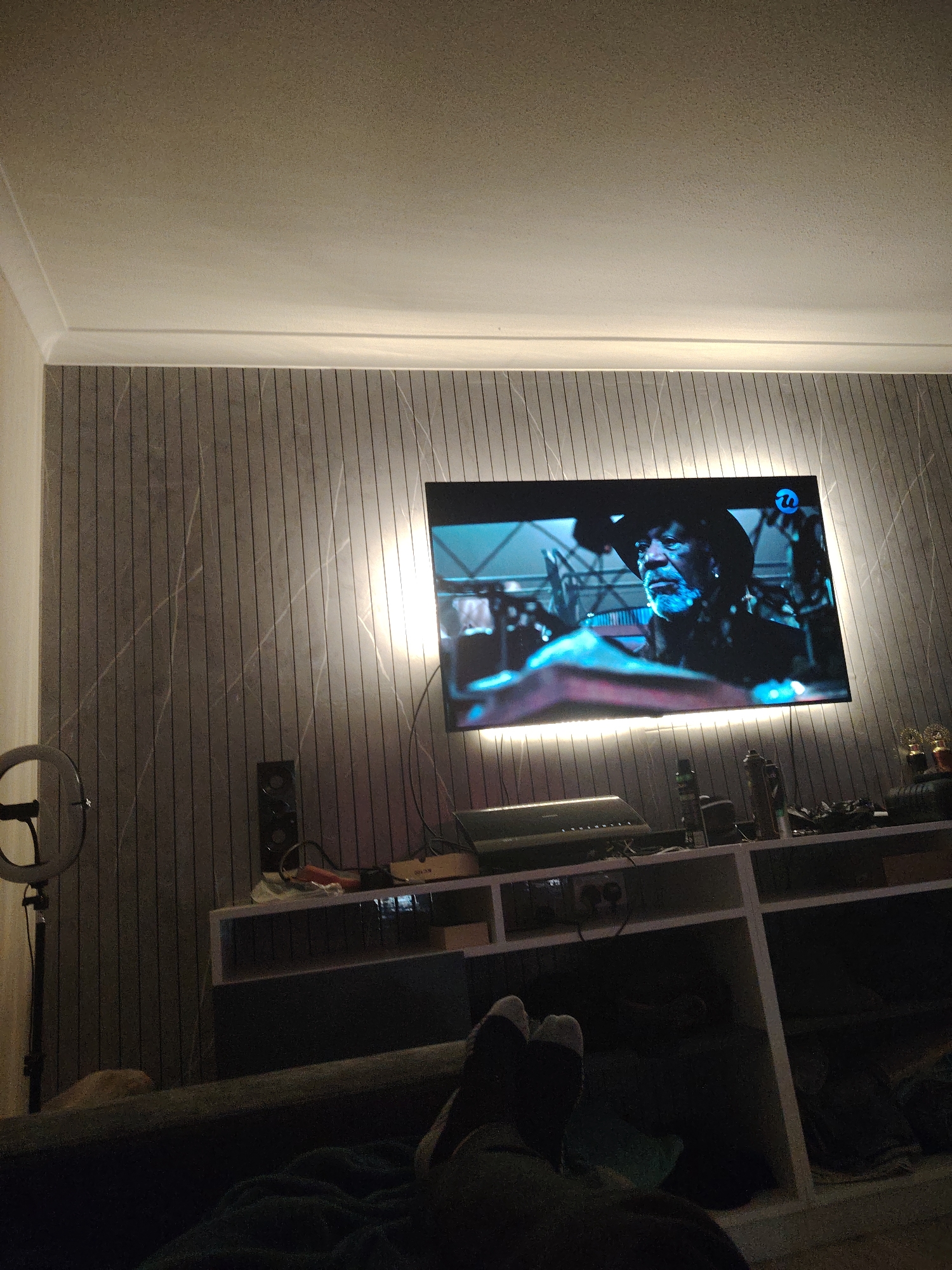 The image depicts a living room setting, with a large television screen displaying a close-up of a man's face wearing a hat and a beard. The television is mounted on a wall that has a vertical paneled design with marble-like veins. Ambient lighting illuminates the wall from behind the television. Below the television, there is a white shelving unit containing various objects, including what appears to be a sound system and some bottles. To the left of the television, a ring light on a stand is visible. In the foreground, a person's legs, clad in socks, are resting on what appears to be a sofa or armchair, suggesting a relaxed viewing posture. The overall impression is a contemporary living space designed for entertainment. There is no information in the image to suggest the location is the city of Huntingdonshire, United Kingdom.