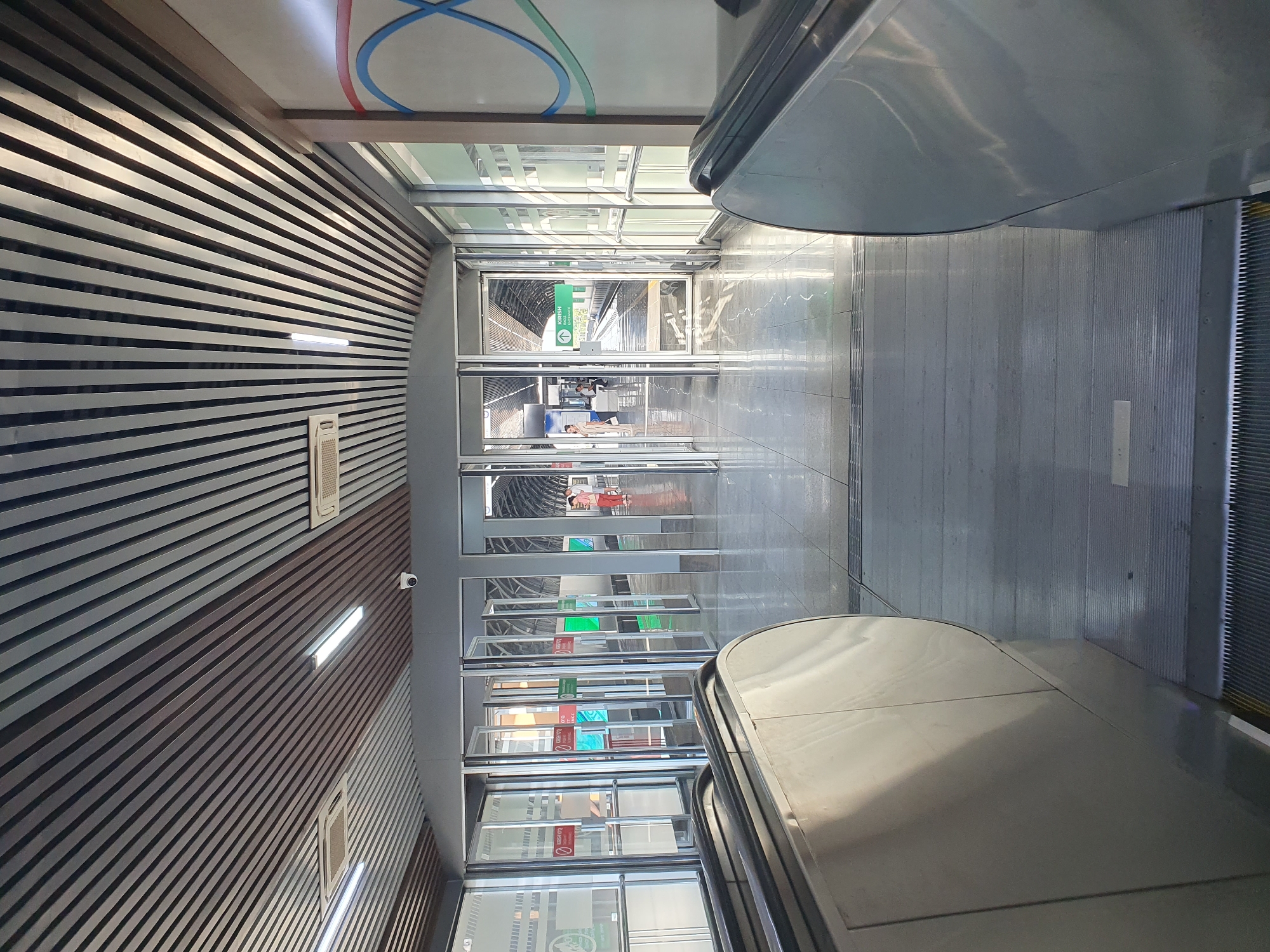 The image shows the interior of a modern train station, likely a metro station, with a perspective looking forward from the base of an escalator. The architecture features a slatted ceiling with integrated lighting and air conditioning units. A security camera is visible on the ceiling. The floor is tiled with a reflective surface. In front, a series of glass-paneled doors lead to platforms where trains are visible. Several people are seen on the platforms, some waiting for trains and others walking. One door has a green sign indicating "KIRISH" and "ENTRANCE" with an arrow, suggesting it is an entrance to the station. The overall environment appears clean and well-maintained, characteristic of urban public transportation infrastructure. The presence of the sign with Cyrillic script, "KIRISH," and the overall design of the station are consistent with modern infrastructure in Tashkent, Uzbekistan.