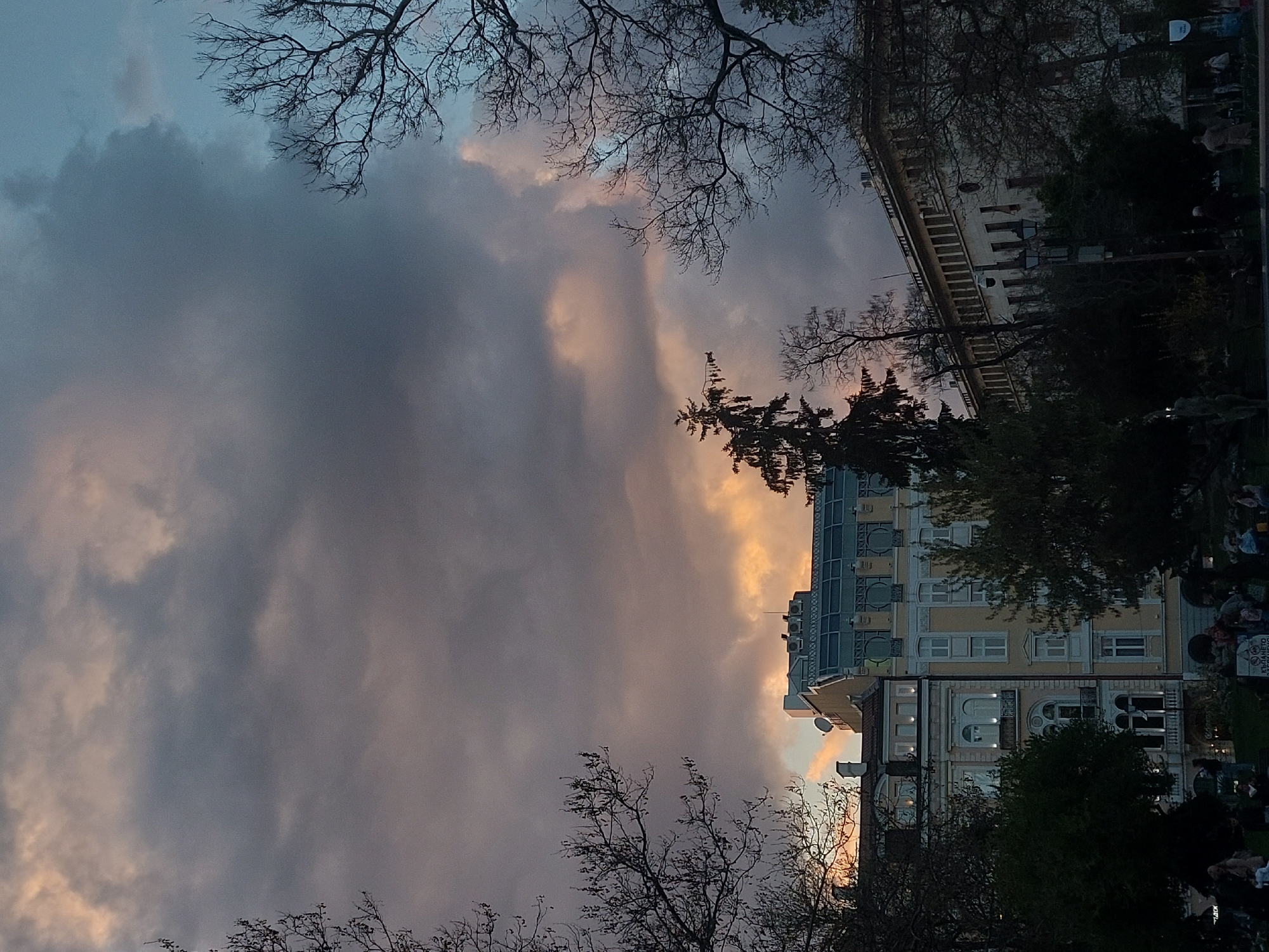 The image captures a twilight scene in an urban setting. The foreground shows bare tree branches silhouetted against a dramatic sky filled with dark, ominous clouds interspersed with patches of peach-colored light. In the middle ground, a light-colored building with multiple stories is partially visible, framed by trees. To the right of the building, there are indistinct figures of people in what appears to be a park or public space. The image is oriented sideways. The location is an unknown city.