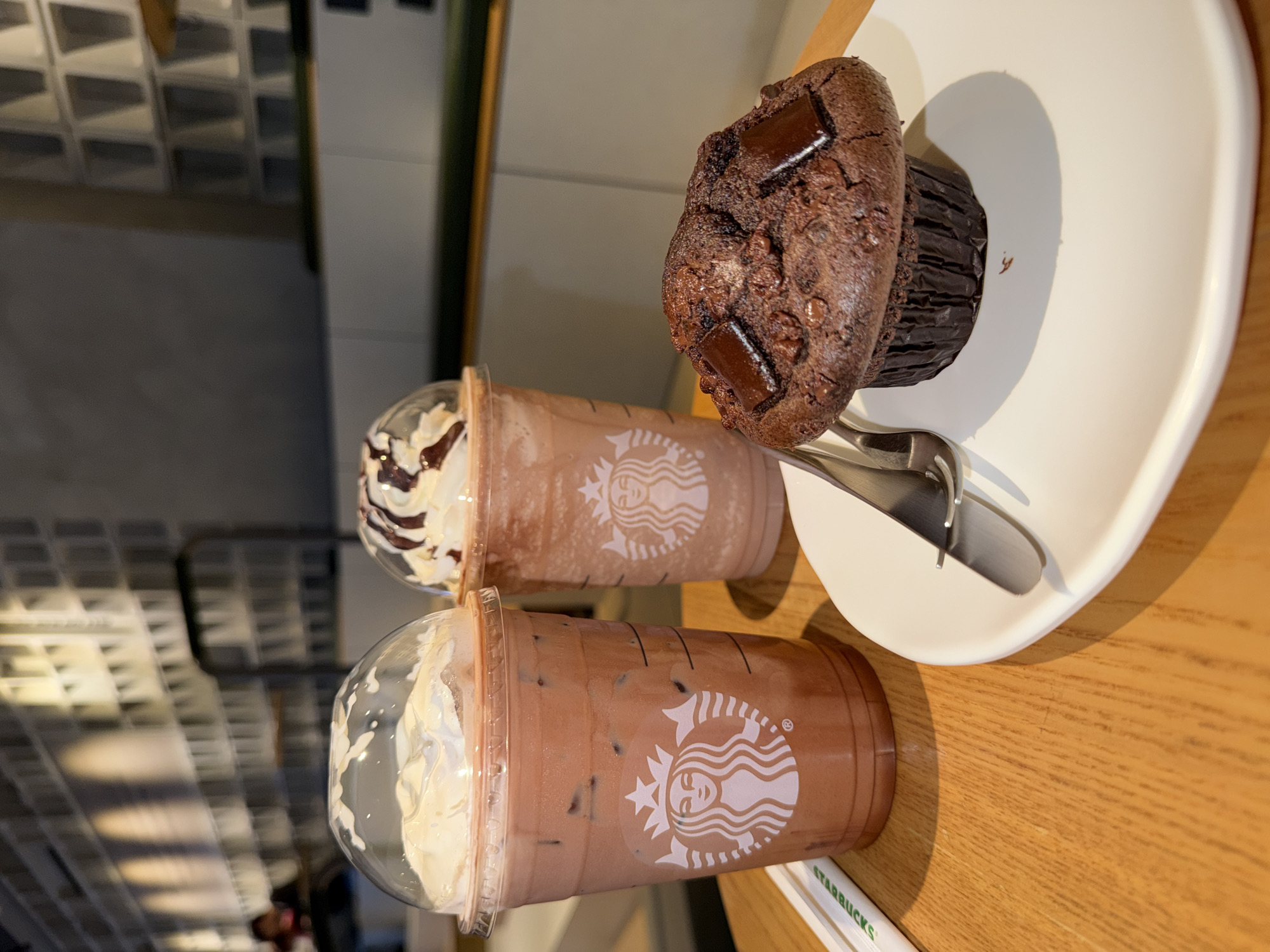 The image depicts a Starbucks café scene, likely in Chongqing High-tech Zone, China, as indicated by the Starbucks logo on the cups. Two clear plastic cups with Starbucks logos are visible, filled with iced chocolate beverages topped with whipped cream. One beverage has chocolate syrup drizzled on top. A chocolate muffin with two chocolate squares on top sits on a small white plate, alongside a fork and knife. The setting appears to be a wooden table within the café, with a partial view of the interior design featuring rectangular wall patterns. The focus is on the food and drinks.