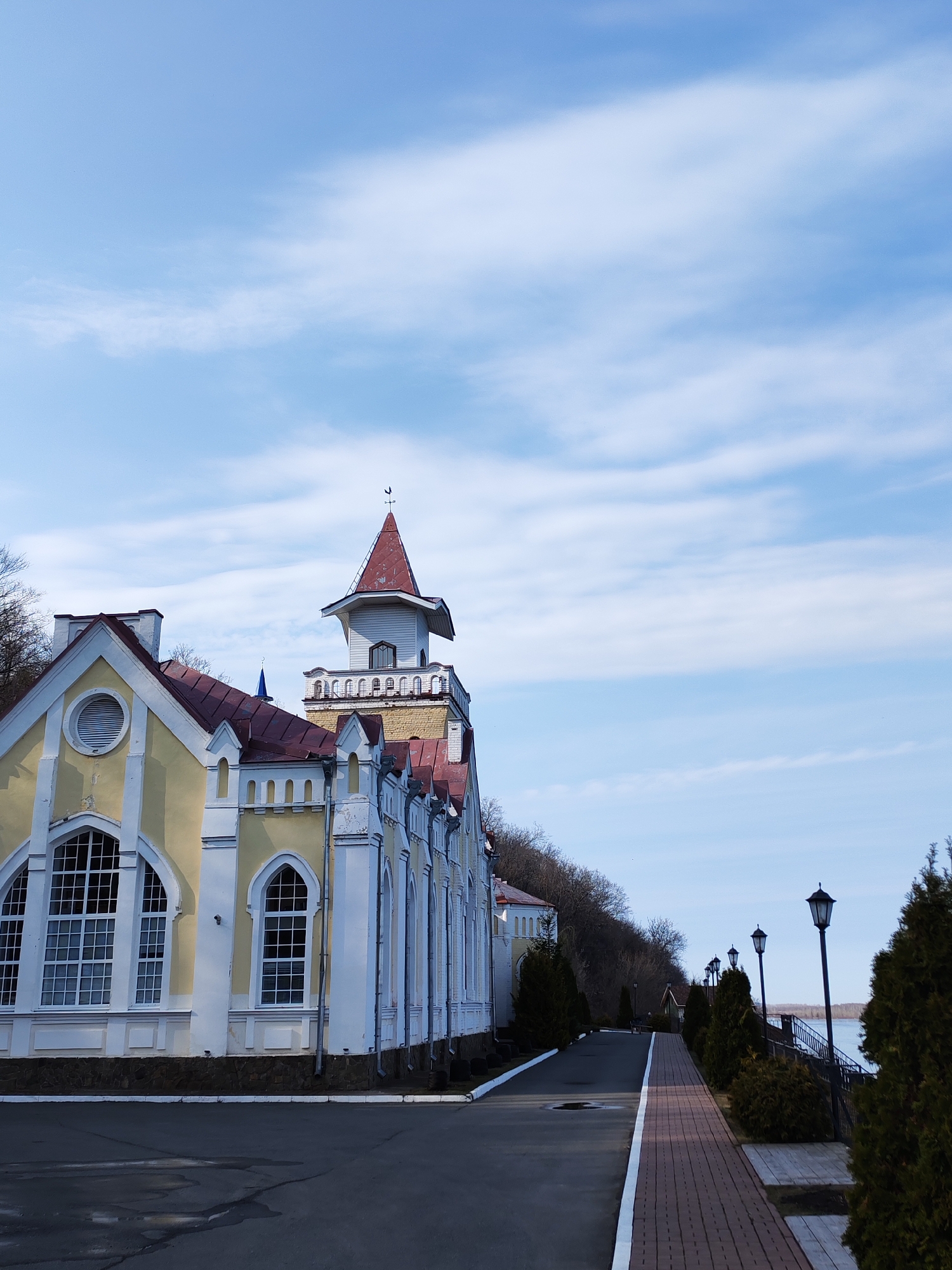 The image shows the Sarapul Hydroelectric Power Station in Sarapul, Russia. The power station, a yellow and white building with a red roof, is on the left side of the image. A road runs along the front of the building, and a brick walkway bordered by evergreen trees and lampposts runs parallel to the road. The sky is blue with scattered white clouds. Trees are visible behind the power station and along the walkway. The river or lake is visible beyond the row of trees. No people are visible in the image.