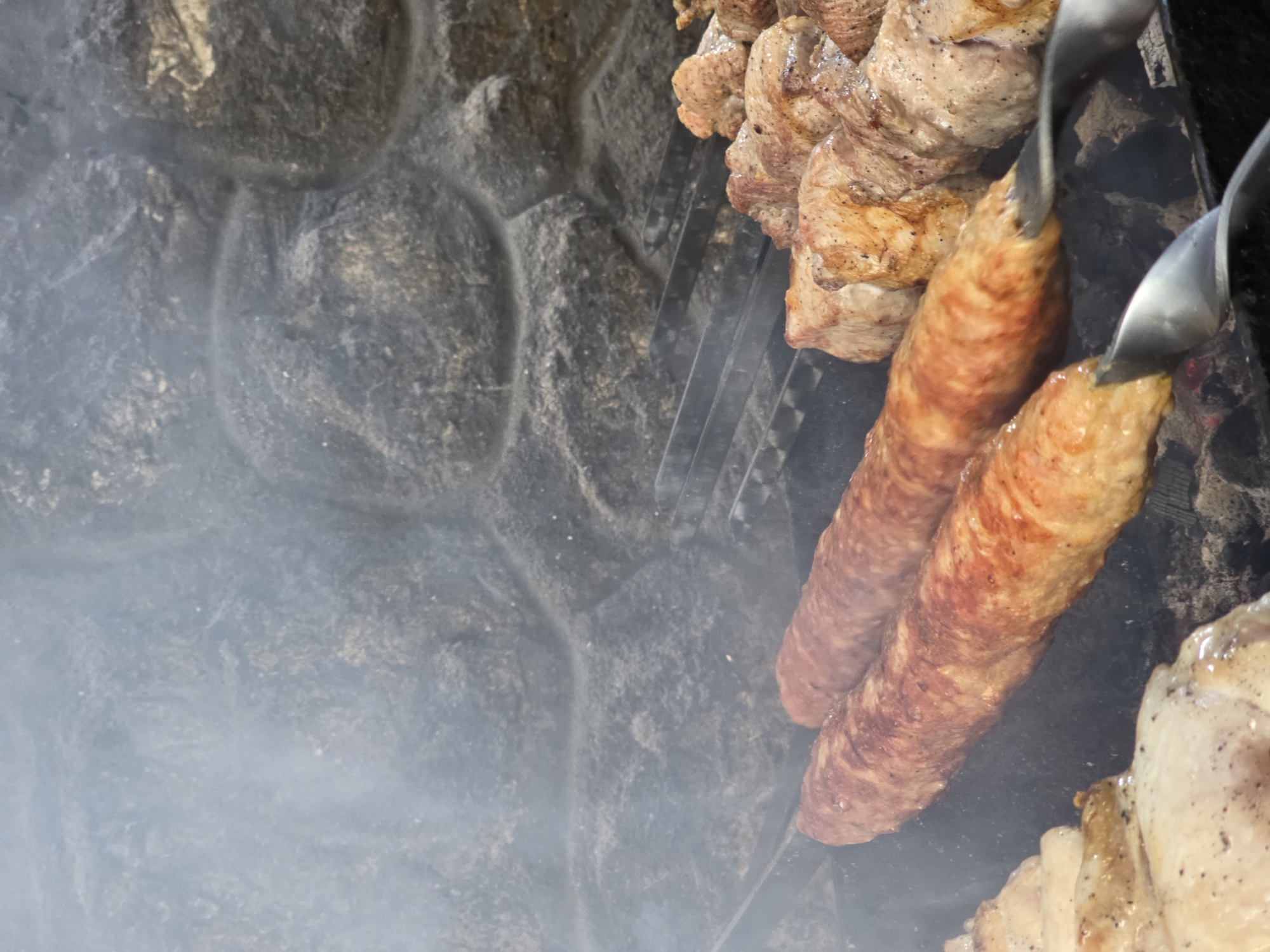 In Pidhorodne, Ukraine, a close-up shot captures two long, sausage-like kebabs cooking on a grill. The kebabs, seasoned and browned, are skewered and positioned prominently in the foreground. Behind them, pieces of cubed meat, also on skewers, are being grilled, their surfaces showing signs of charring and seasoning. Wisps of smoke rise from the grill, indicating active cooking. The background features a rough, stone-like surface, possibly part of an outdoor grill or a rustic wall, adding a natural and earthy element to the scene. The lighting is dim, suggesting it might be evening or an overcast day, with the focus on the food and the cooking process.