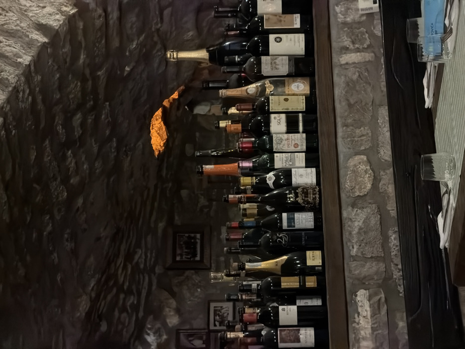 This image captures an interior view of a restaurant or wine cellar located in Morinj, Montenegro. The background and upper portions of the scene are dominated by an irregular, dark-toned natural stone wall and ceiling. An opening in the upper right section of this stone structure emits a distinct orange light, illuminating the immediate area.
A prominent collection of approximately 30 to 40 assorted wine and champagne bottles is displayed in a single row on a dark wooden shelf, spanning horizontally across the mid-ground. The bottles vary in color, size, and label design; some partially visible labels include "KRUG," "SOLAIS," "PETRUS," and "ORNELIA." To the left of the bottle display, several framed pictures are affixed to the stone wall.
Below the main bottle shelf, a section of lighter-toned, more regularly coursed stone masonry is visible, topped by a darker wooden beam. In the foreground, a dining table is partially set, covered with a light-colored checkered tablecloth. A clear, textured drinking glass is positioned on the table, accompanied by a folded white napkin containing a fork. Another similar drinking glass is partially visible on the far right. No individuals are present in the scene.